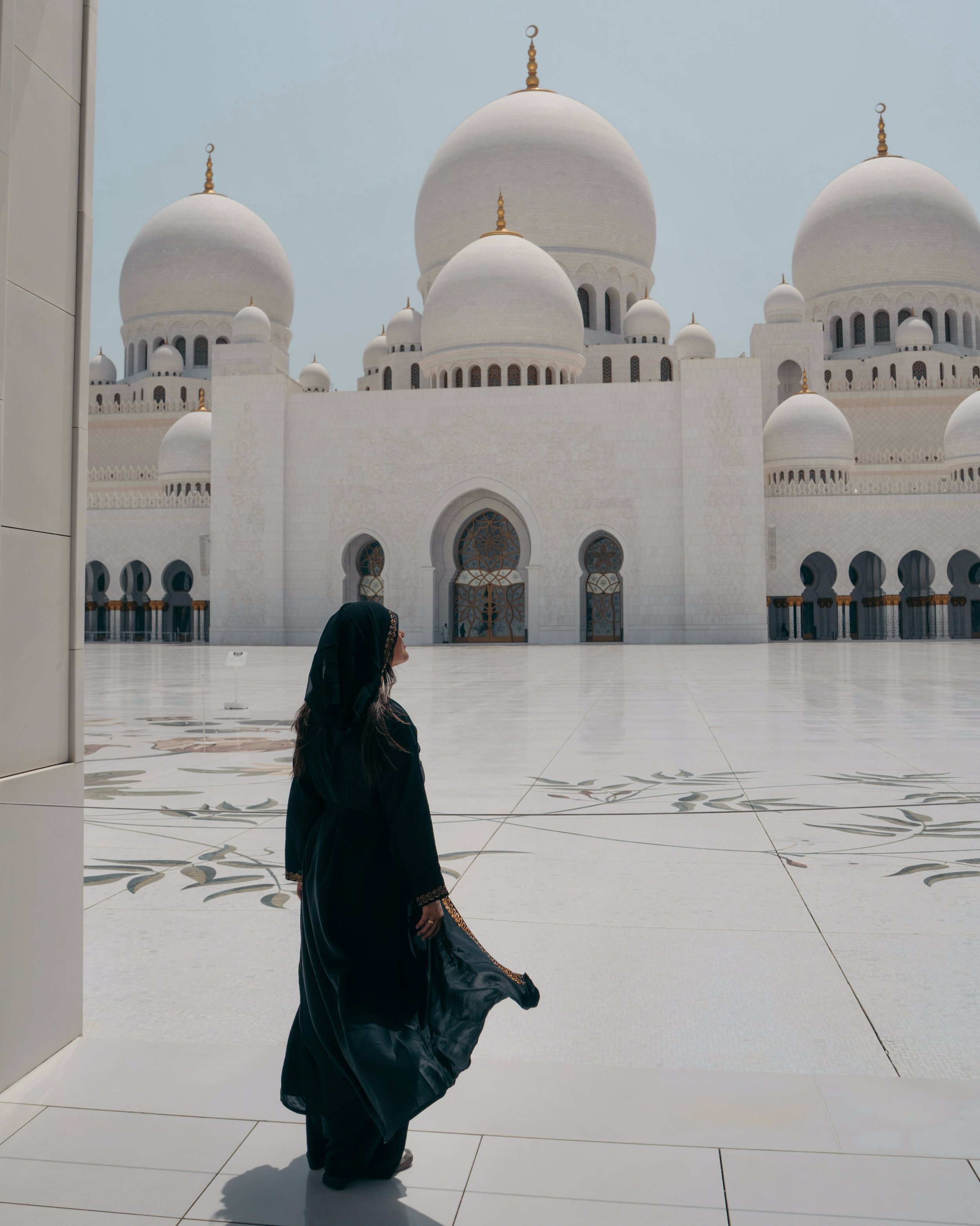 A woman in a black abaya standing in front of the Sheikh Zayed Grand Mosque's white marble exterior and domes.