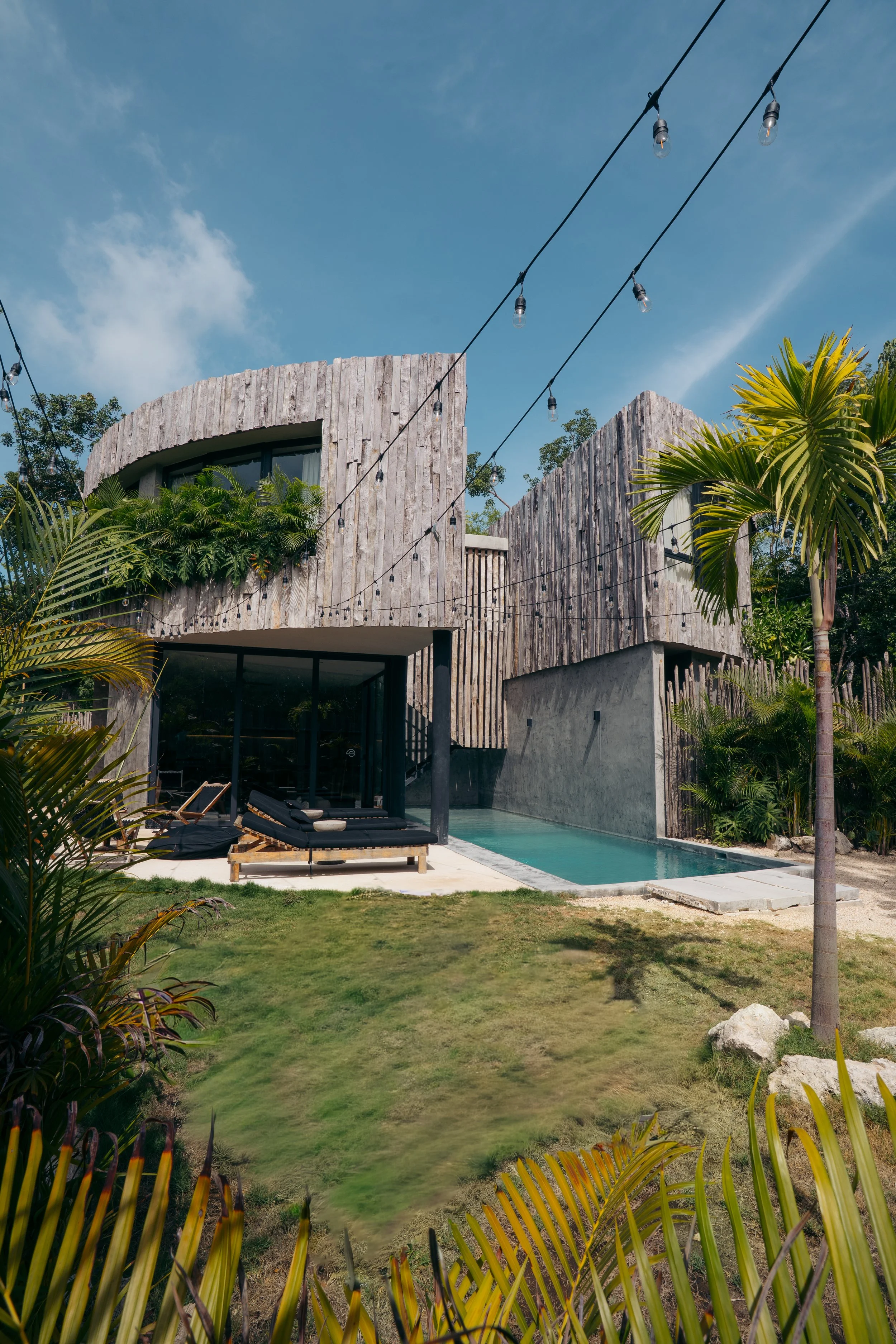 Modern house with a curved wooden exterior, outdoor seating area, a pool, surrounded by lush tropical plants and trees, with string lights hanging overhead, under a partly cloudy sky.