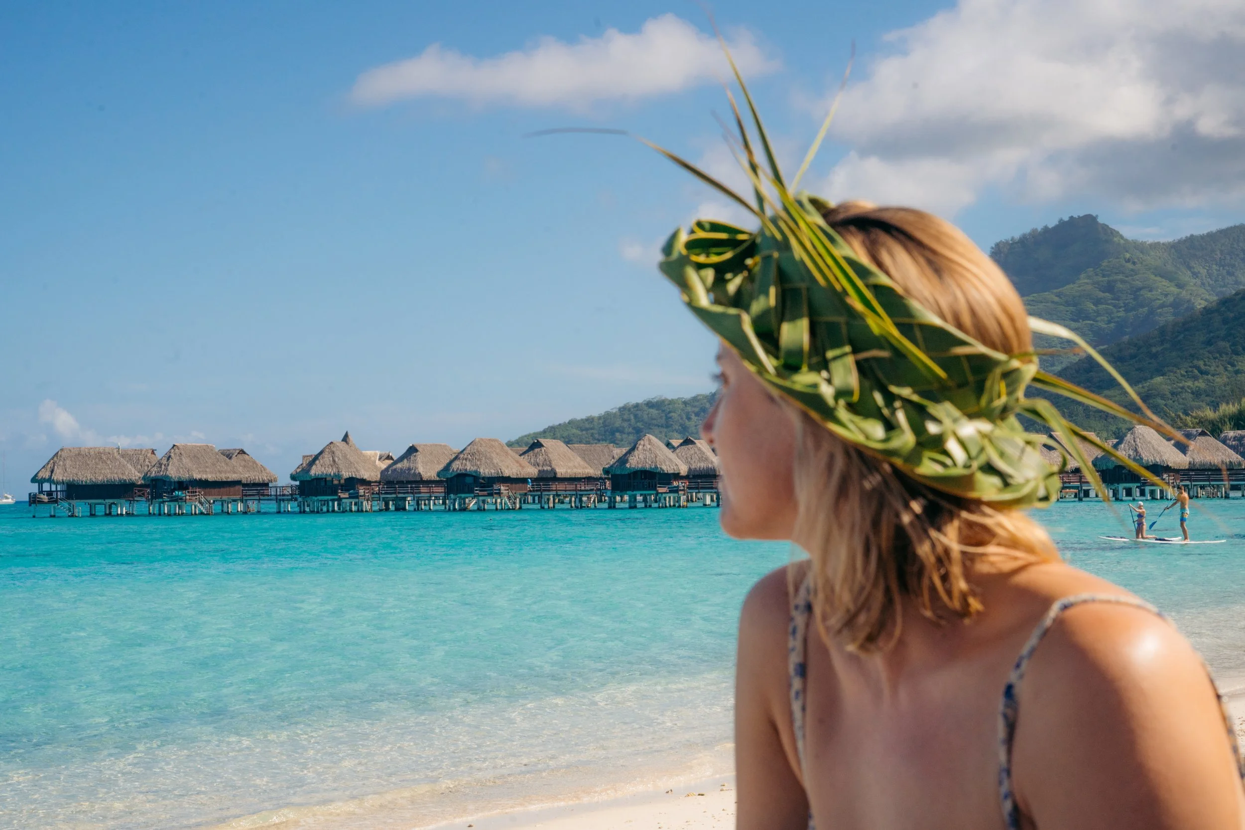 A woman with blonde hair wearing a headpiece made of green palm leaves, standing on a beach with overwater bavilions and a mountain in the background.
