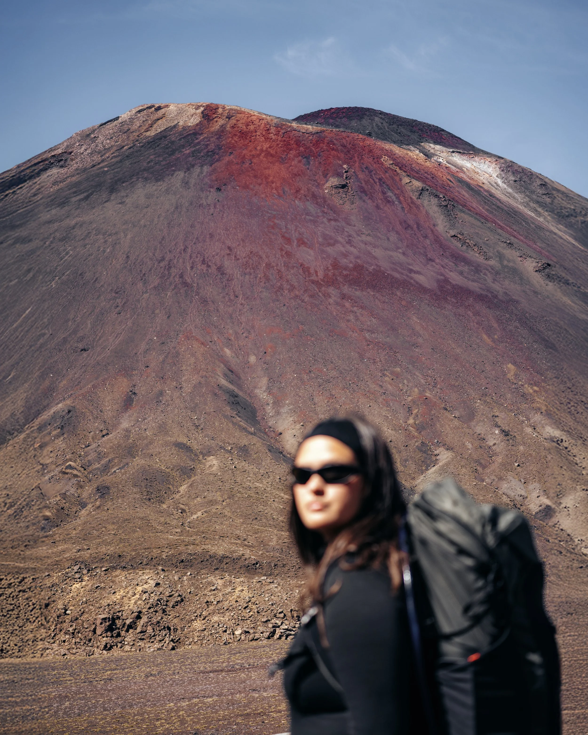 A person with dark hair, wearing sunglasses and a black jacket, is falling or leaning forward in front of a volcanic mountain with reddish and brownish terrain and a blue sky background.
