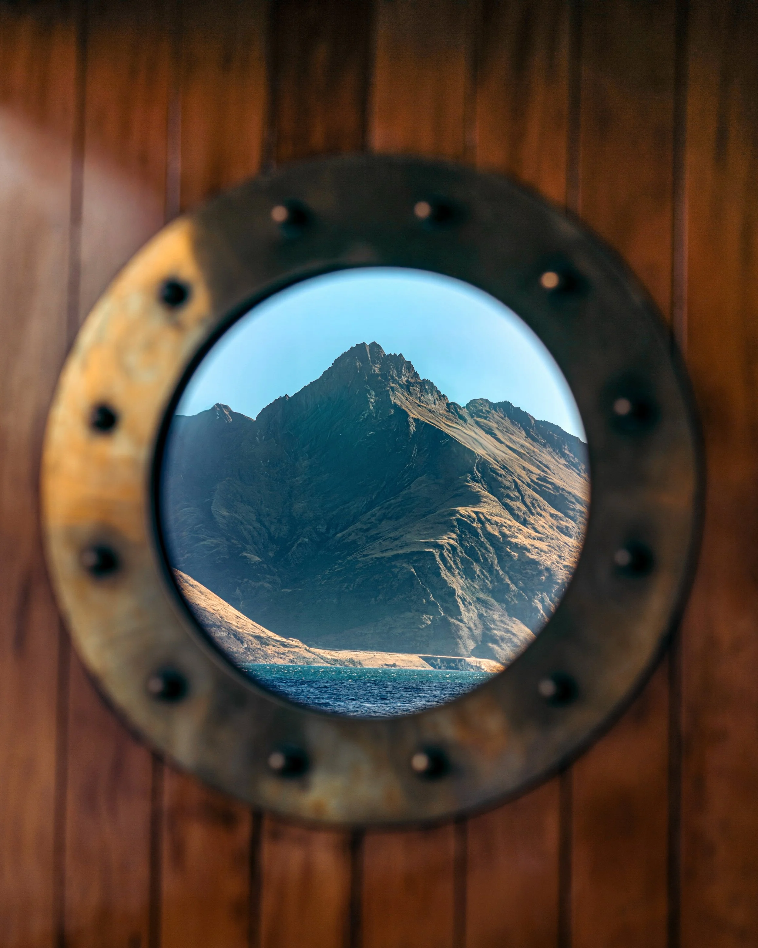 A mountain range with rugged peaks viewed through a round porthole with a metallic frame, mounted on a wooden wall.