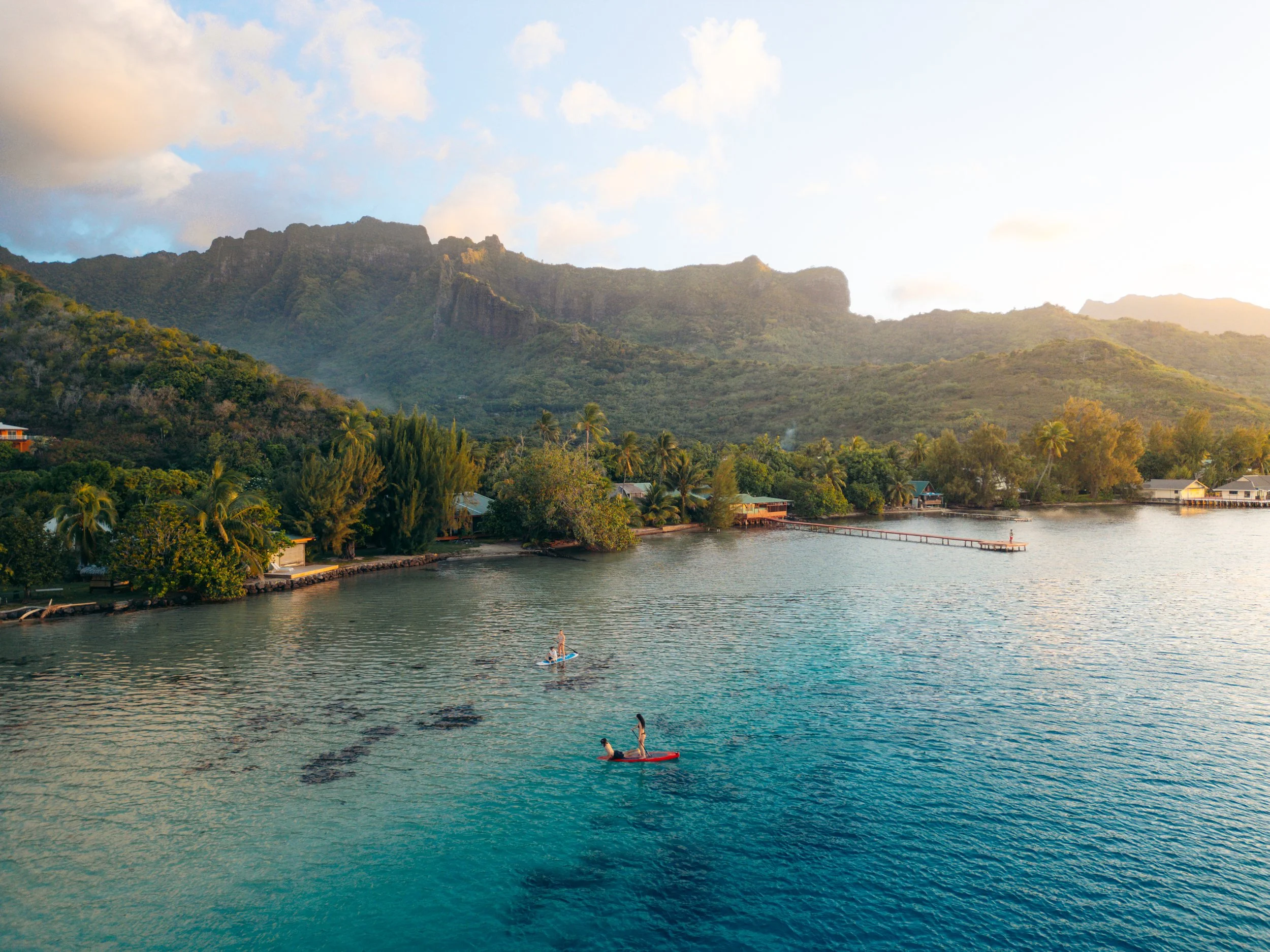 People paddleboarding on calm water near lush green trees and a mountain with rocky cliffs in the background, under a partly cloudy sky.