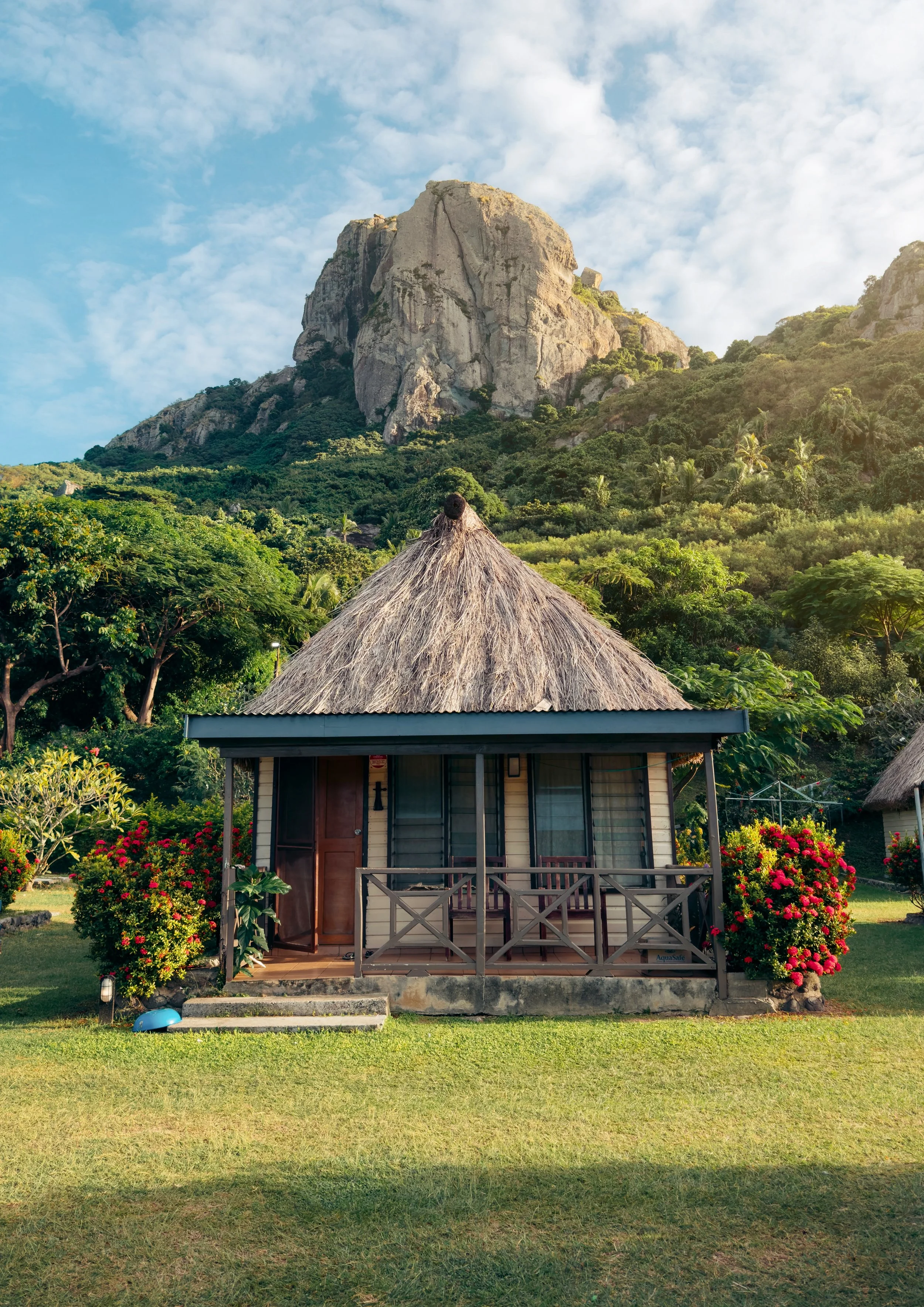 A small house with a thatched roof in a lush green garden, with colorful flowers and shrubs in the foreground, and a rocky mountain and blue sky with clouds in the background.