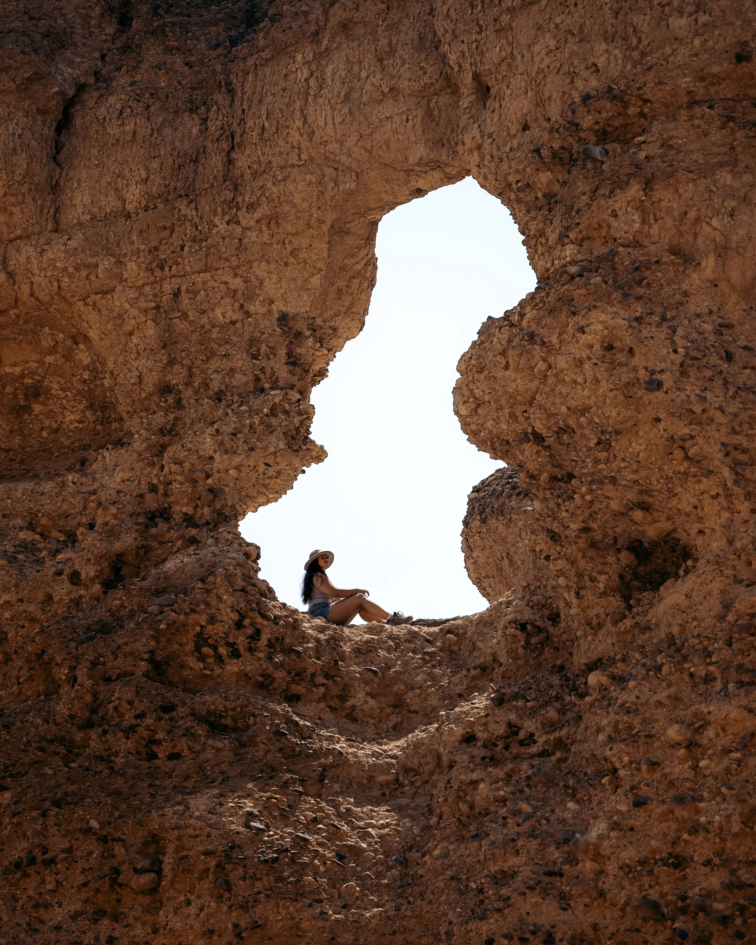 A woman sitting at the edge of a large hole in a rock formation, wearing a hat and looking upward, with clear sky visible through the hole.