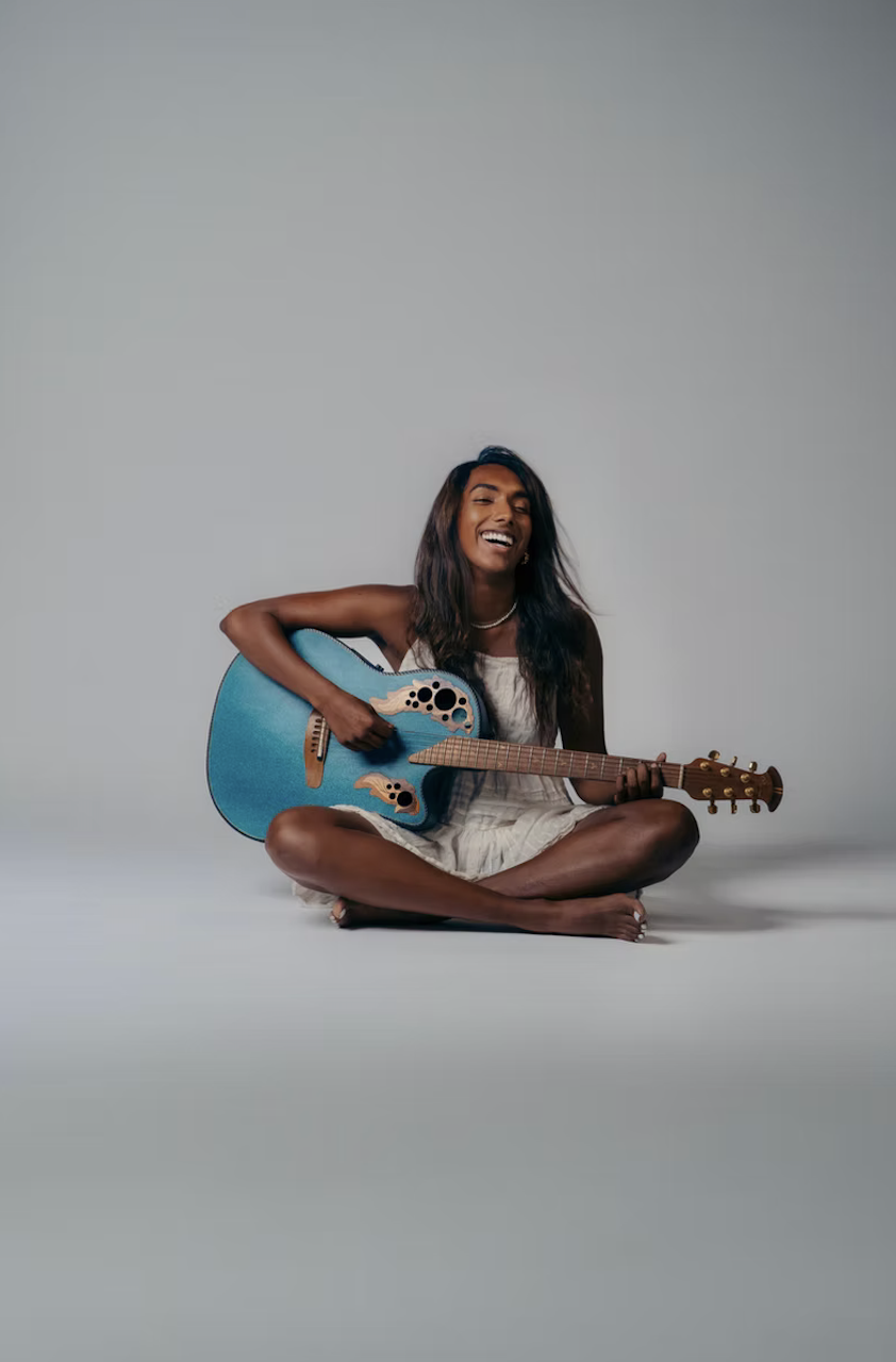 A woman sitting cross-legged on the floor, smiling while playing a blue acoustic guitar, against a plain gray background.