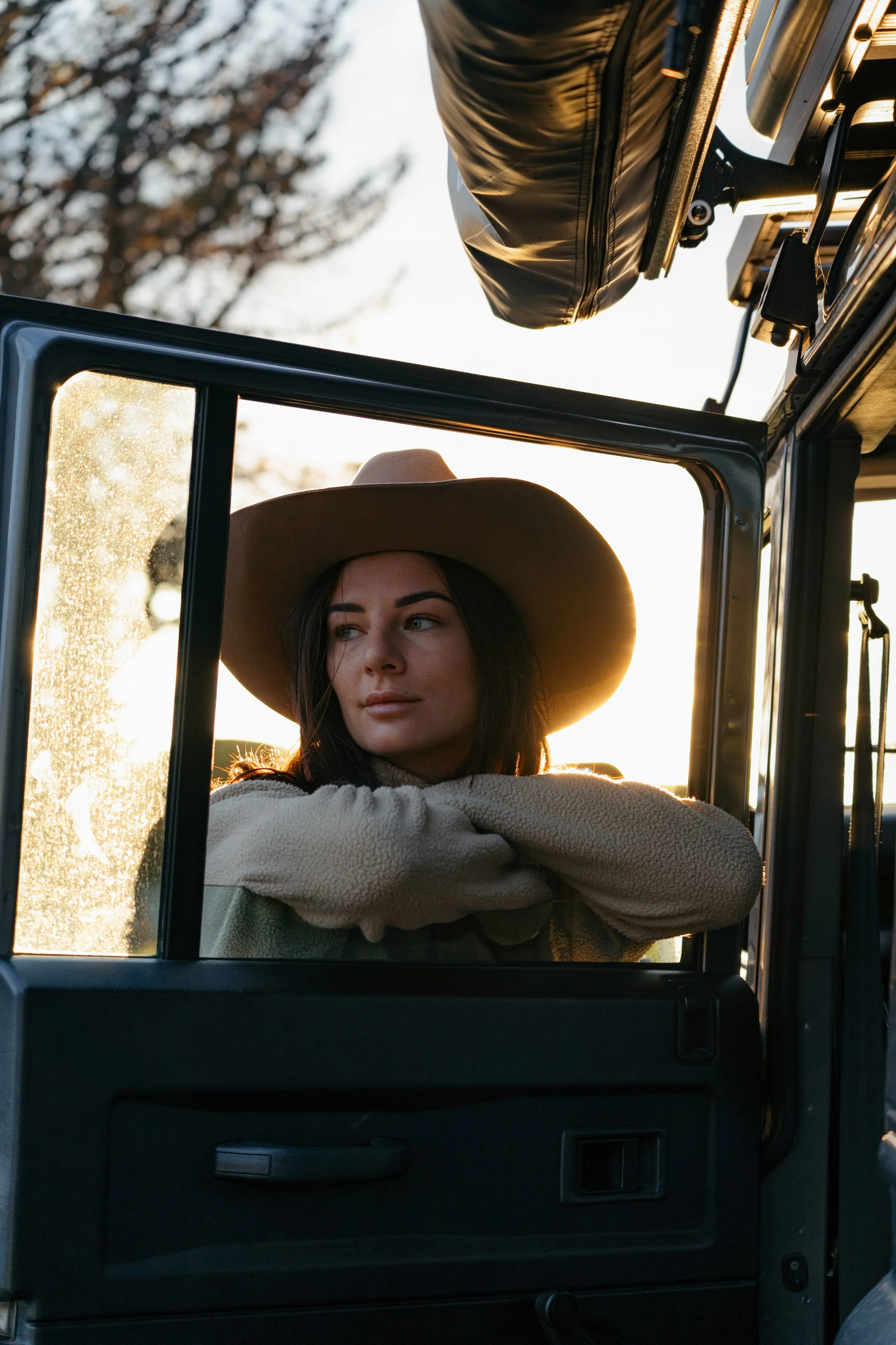 A woman with brown hair and a wide-brimmed hat sitting inside a vehicle, possibly a jeep or similar, with her arms crossed on the window ledge. The sun is setting or rising, casting a warm glow through the window, with trees visible in the background