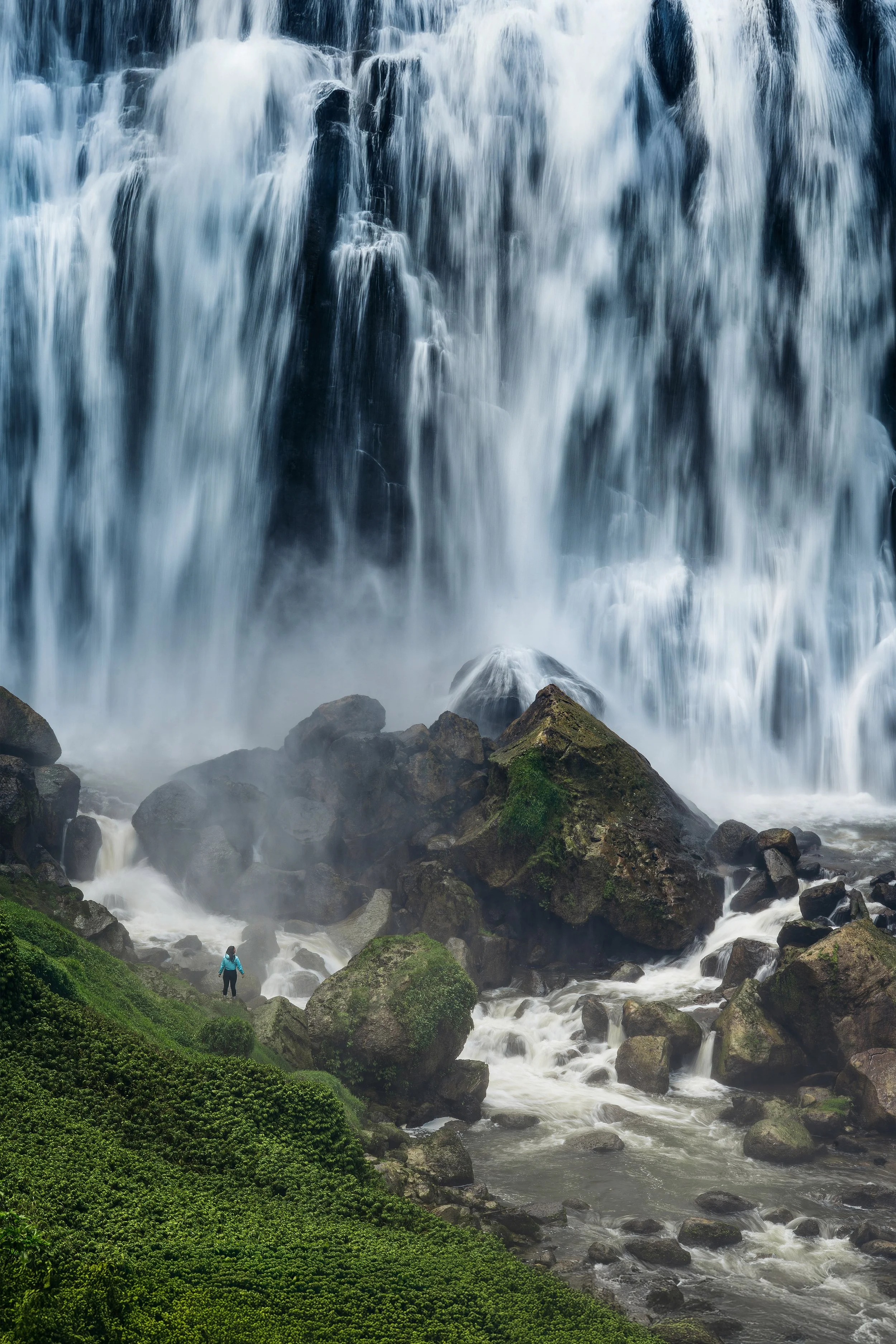 A person in a blue jacket standing on rocks in front of a large waterfall with water cascading down over rocks, surrounded by green moss and plants.