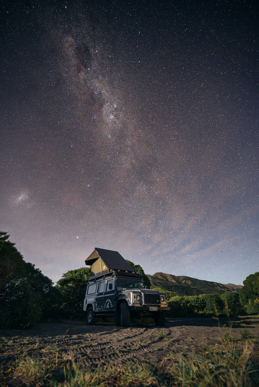 A rugged vehicle with a rooftop tent parked on a dirt path in a remote area under a starry night sky, with the Milky Way galaxy visible.