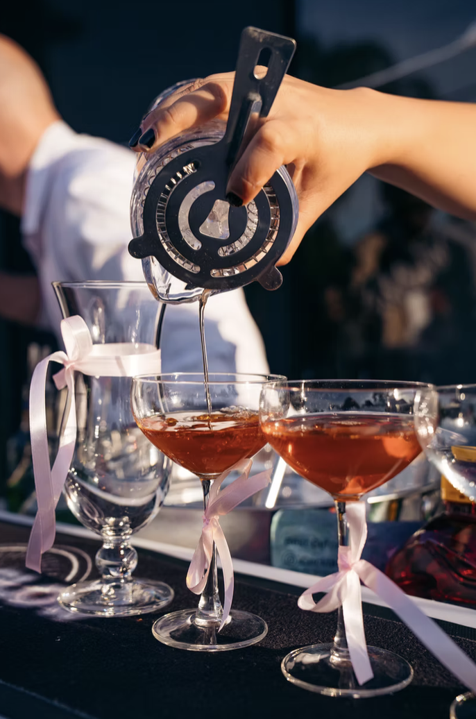 Person pouring a drink into cocktail glasses decorated with pink ribbons at an outdoor celebration.