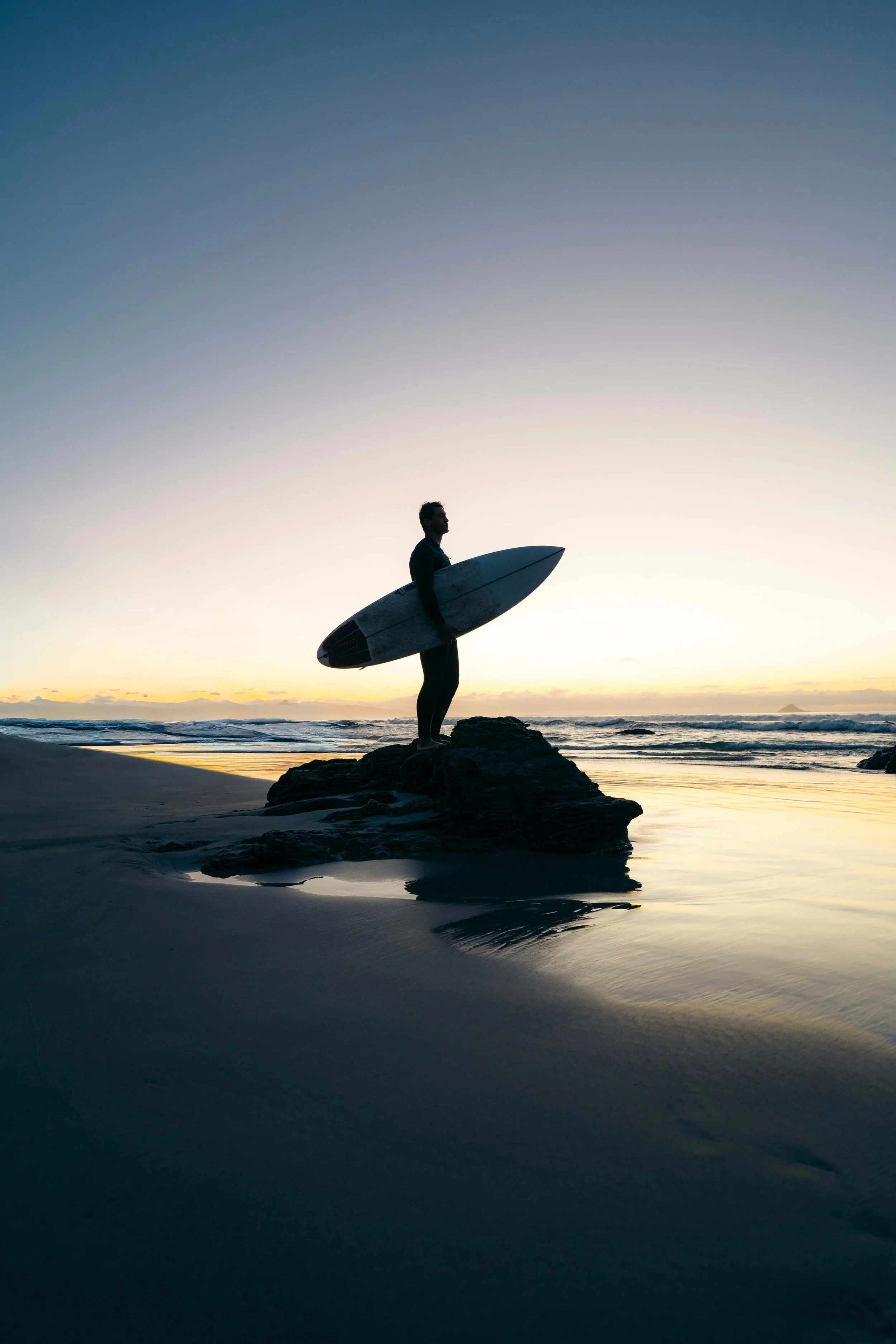 Person holding a surfboard standing on rocks at the beach during sunset, with calm ocean waves and a clear sky.