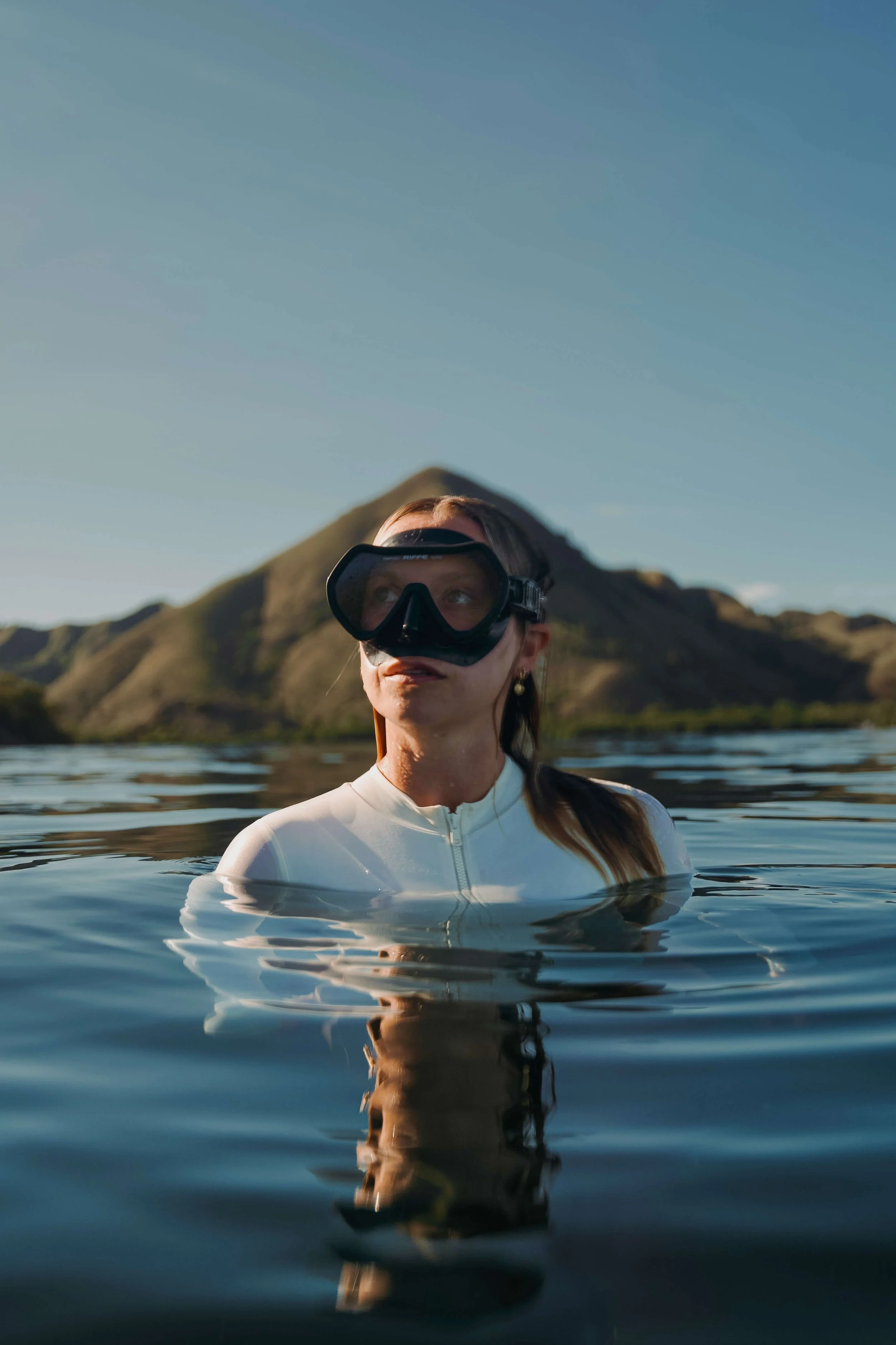 A woman wearing a white wetsuit and a diving mask is standing in the water, with mountains and a clear sky in the background.