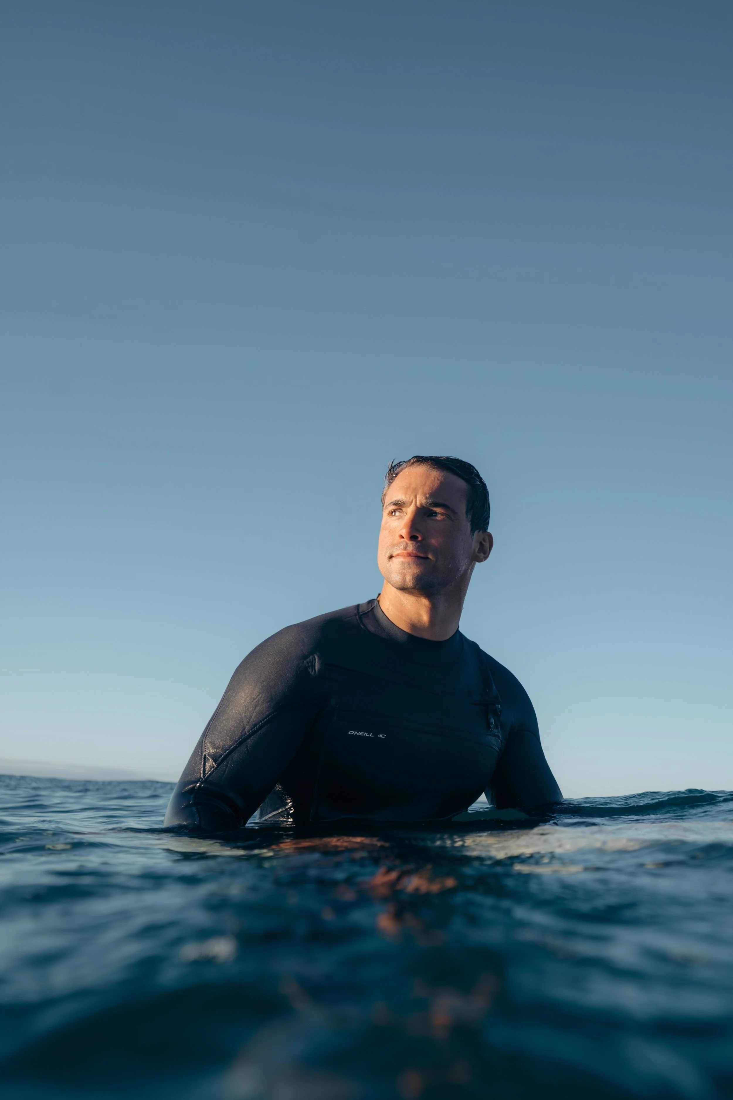 A man in a black wetsuit sitting in the water outdoors, with a clear blue sky above and calm water, looking to the side.