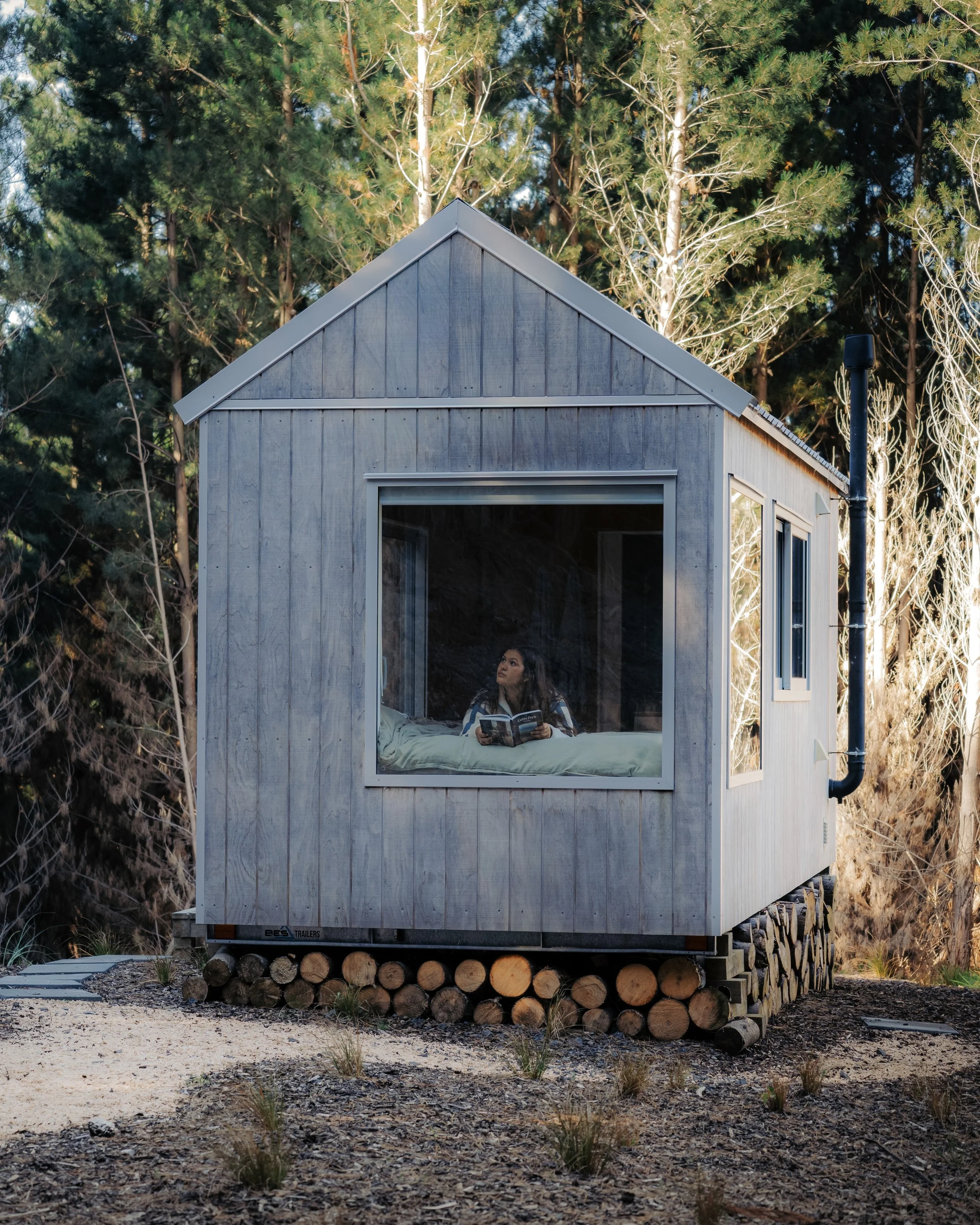 A woman sitting inside a small, modern tiny house with large window, reading a book, with forest trees behind it.