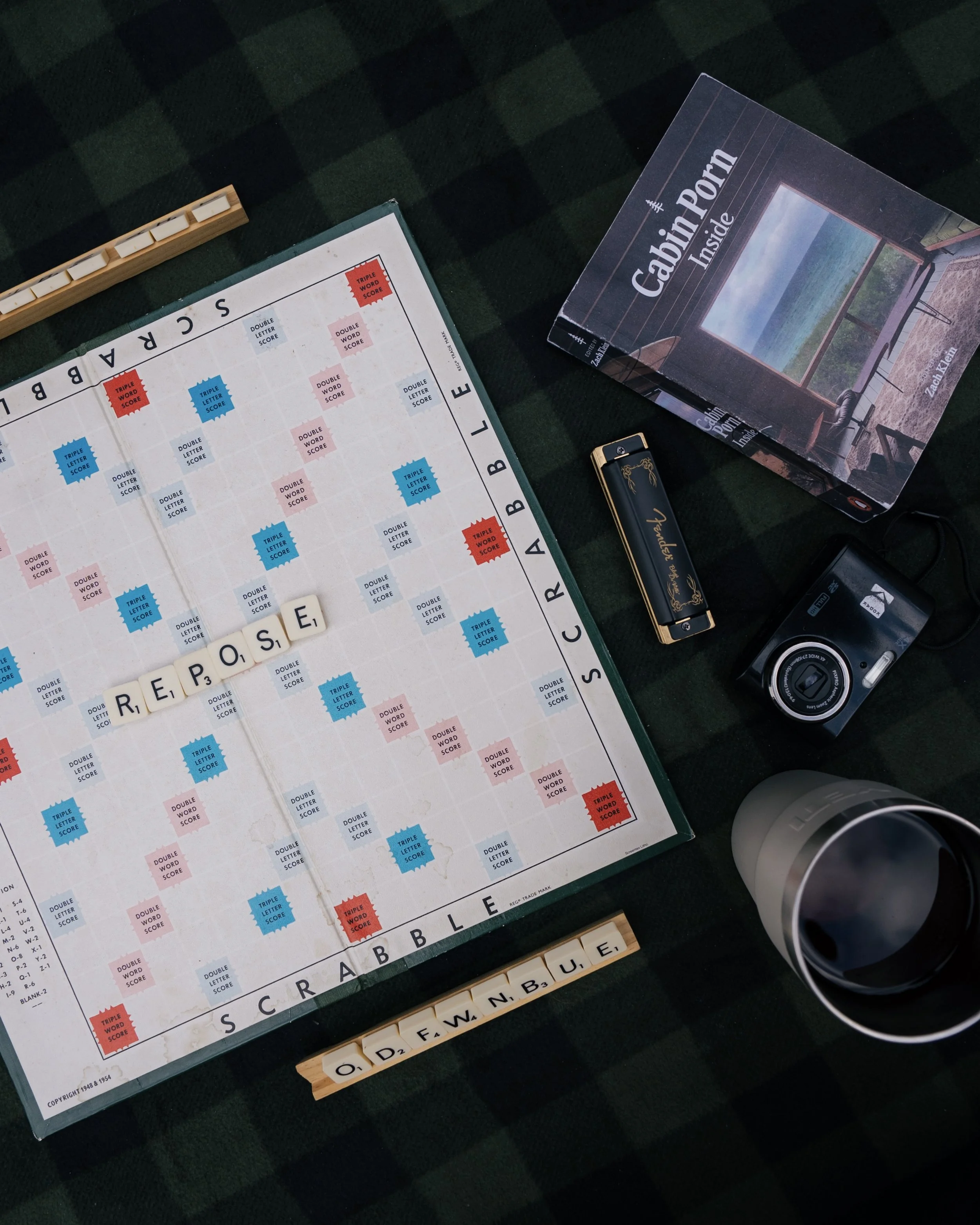 Scrabble game with letter tiles spell out 'RESPONSE' on the game board, a box labeled 'Cabin Porch Inside' showing a picture of a porch with a scenic view, a vape device, a camera, and two cups on a dark green checkered surface.