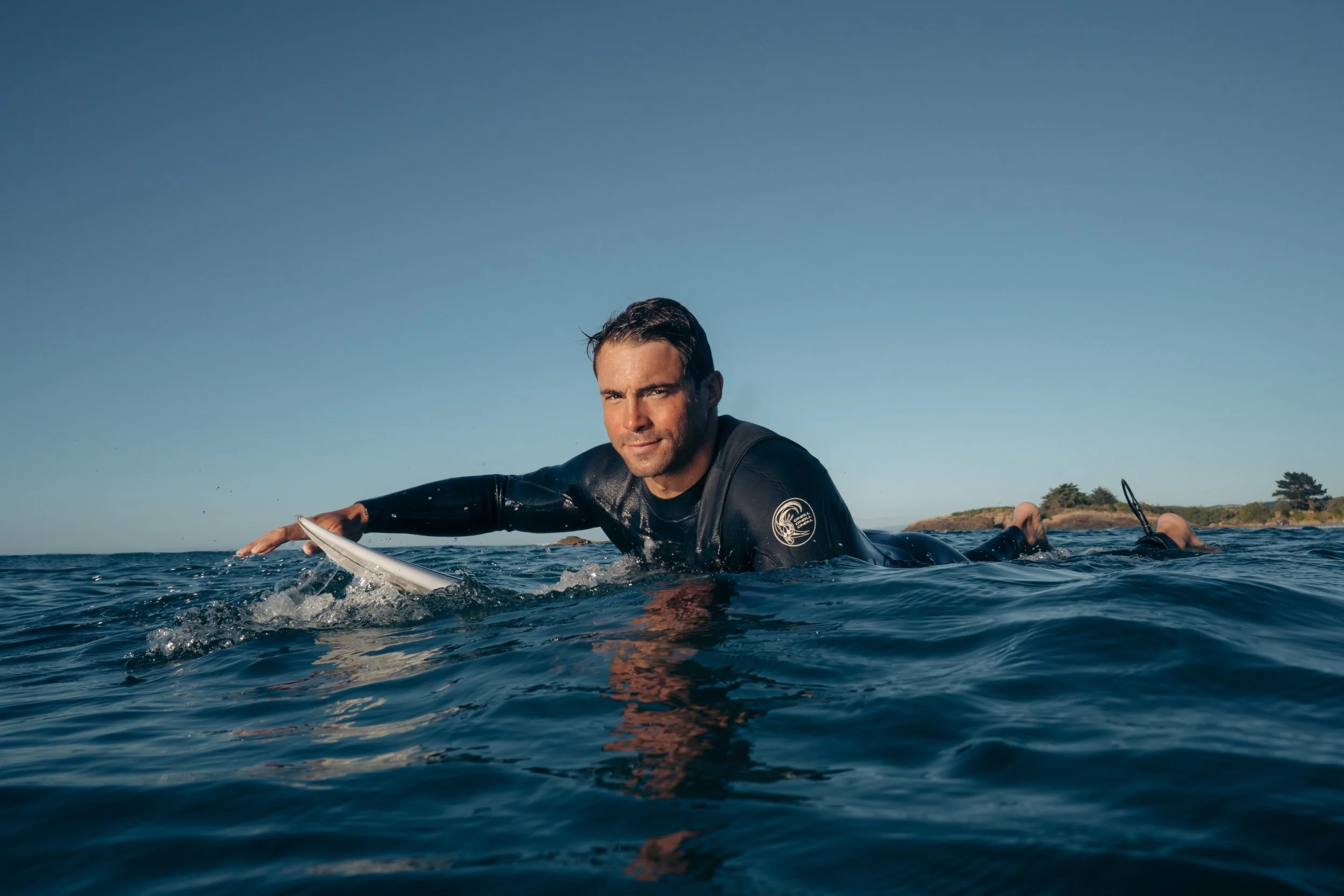 A man in a wetsuit paddling on a surfboard in the water with a clear sky and distant shoreline in the background.