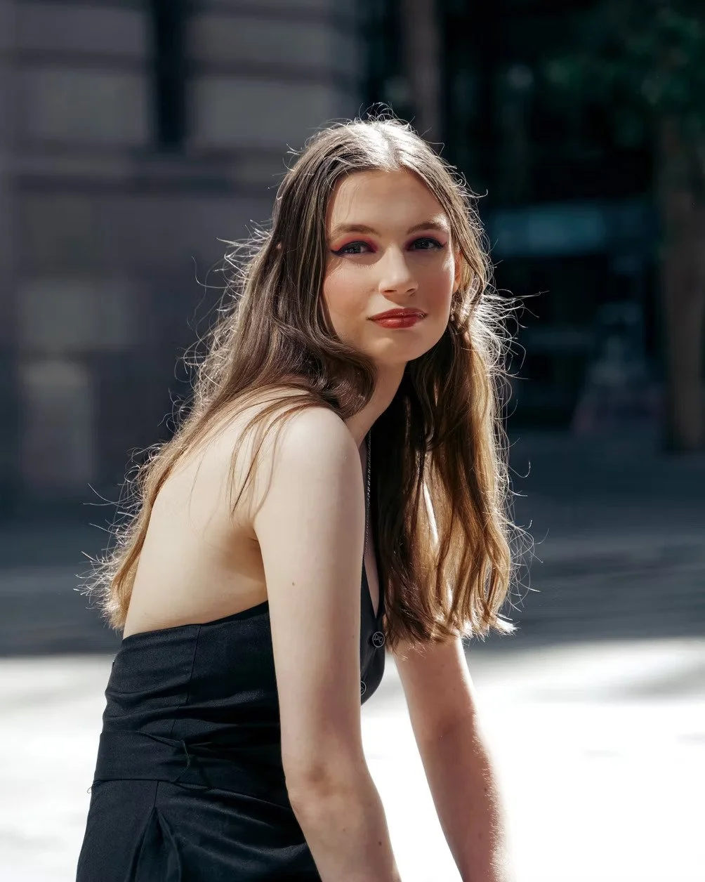 A young woman with long, wavy brown hair, wearing a black sleeveless top, sitting outdoors, looking at the camera with a slight smile.
