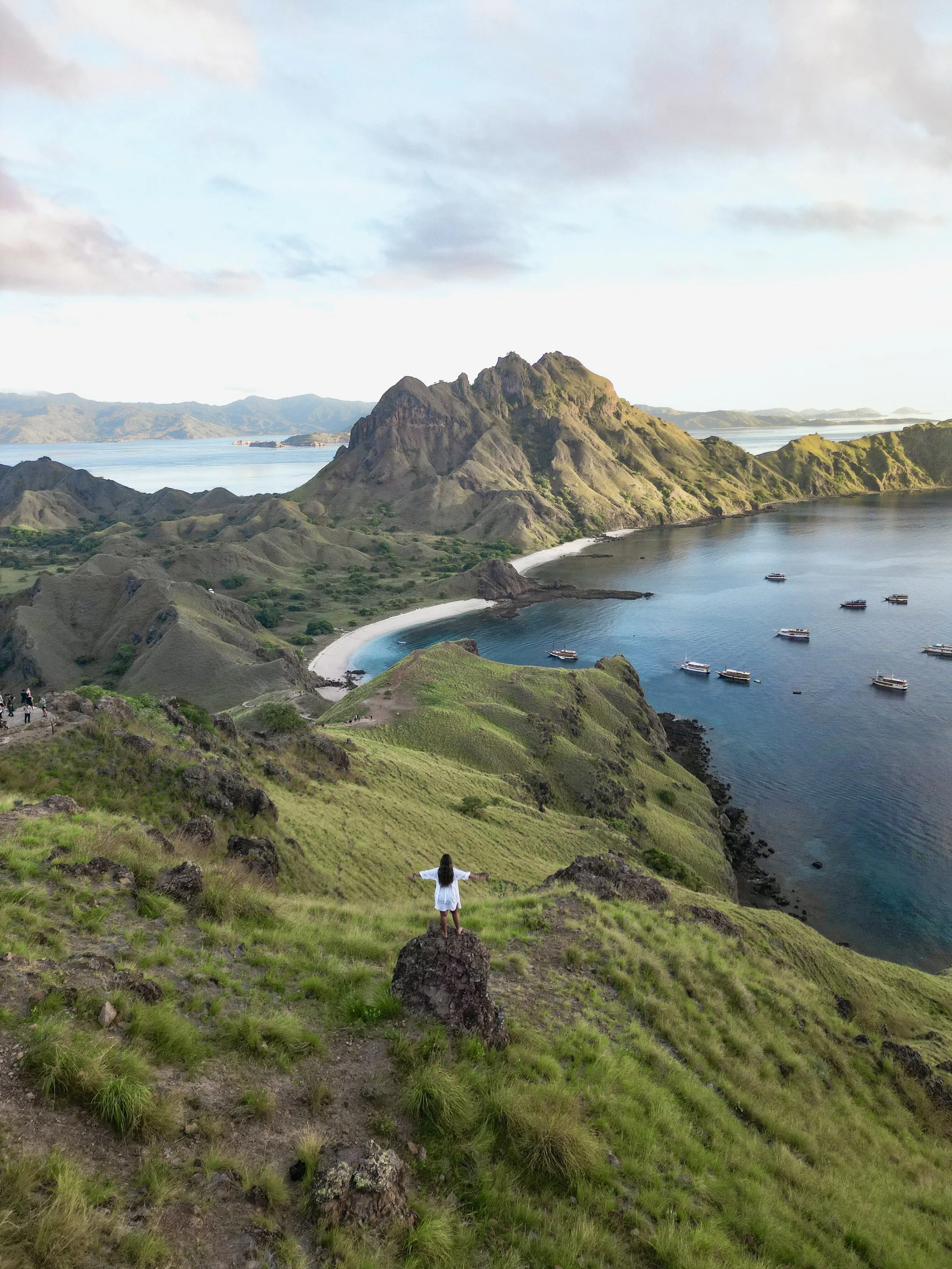 A woman stands with arms outstretched on a grassy hill overlooking a bay with boats, mountains, and a cloudy sky.