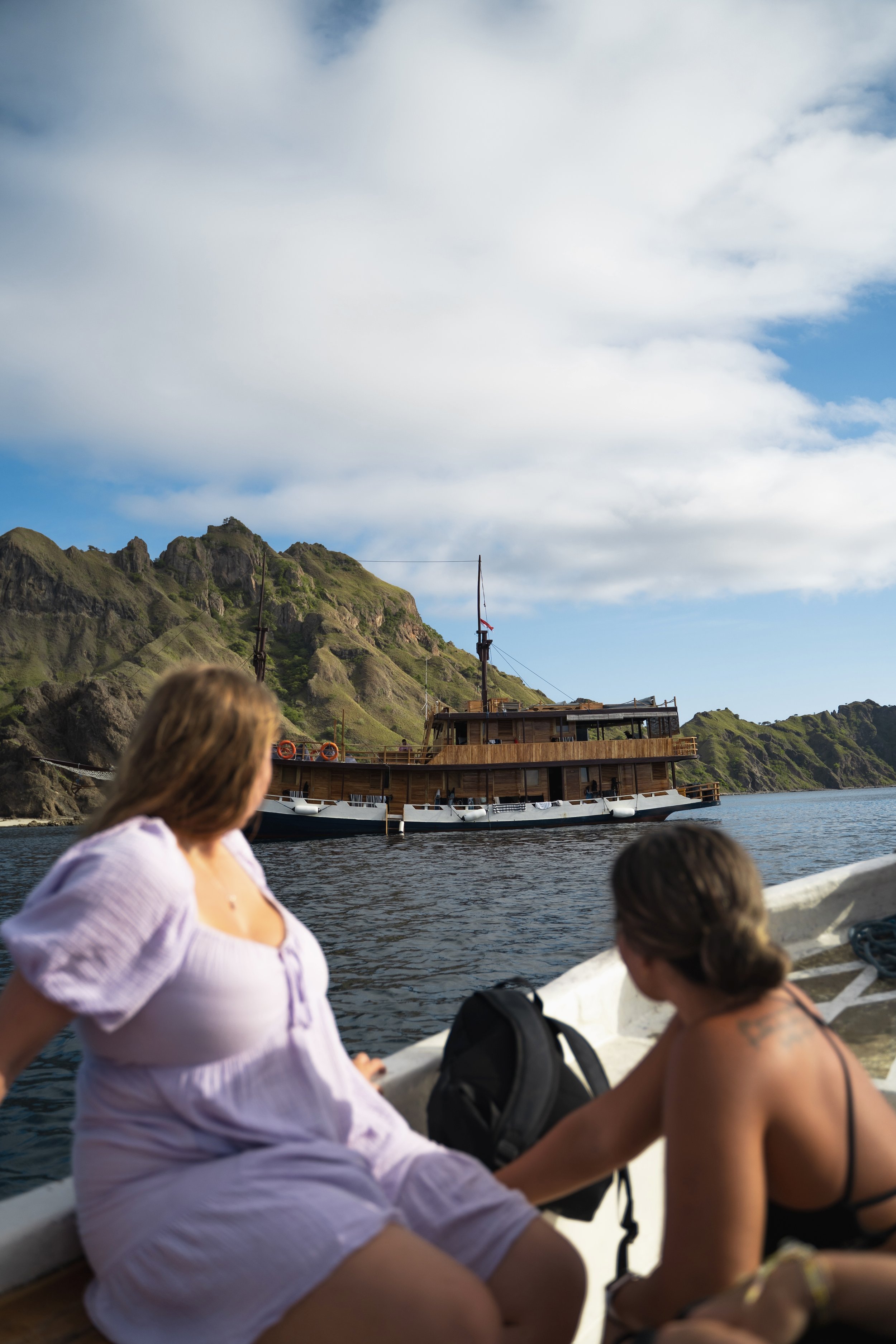 Multiple people sitting on a boat near a coastal area with green hills and a house in the background, under cloudy sky.