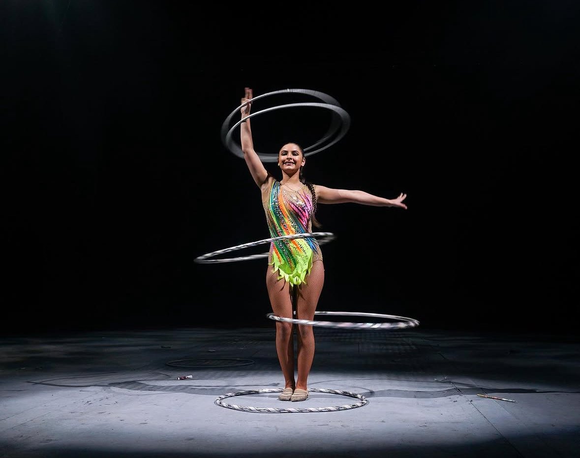 A female gymnast performing a hula hoop routine on stage, balanced with multiple hoops around her body and a bright smile, wearing a colorful leotard against a black background.