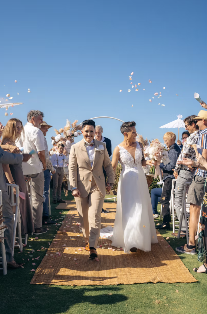A couple, one in a beige suit and the other in a white wedding dress, walking down an outdoor aisle at their wedding, surrounded by guests and throwing flower petals, under a clear blue sky.