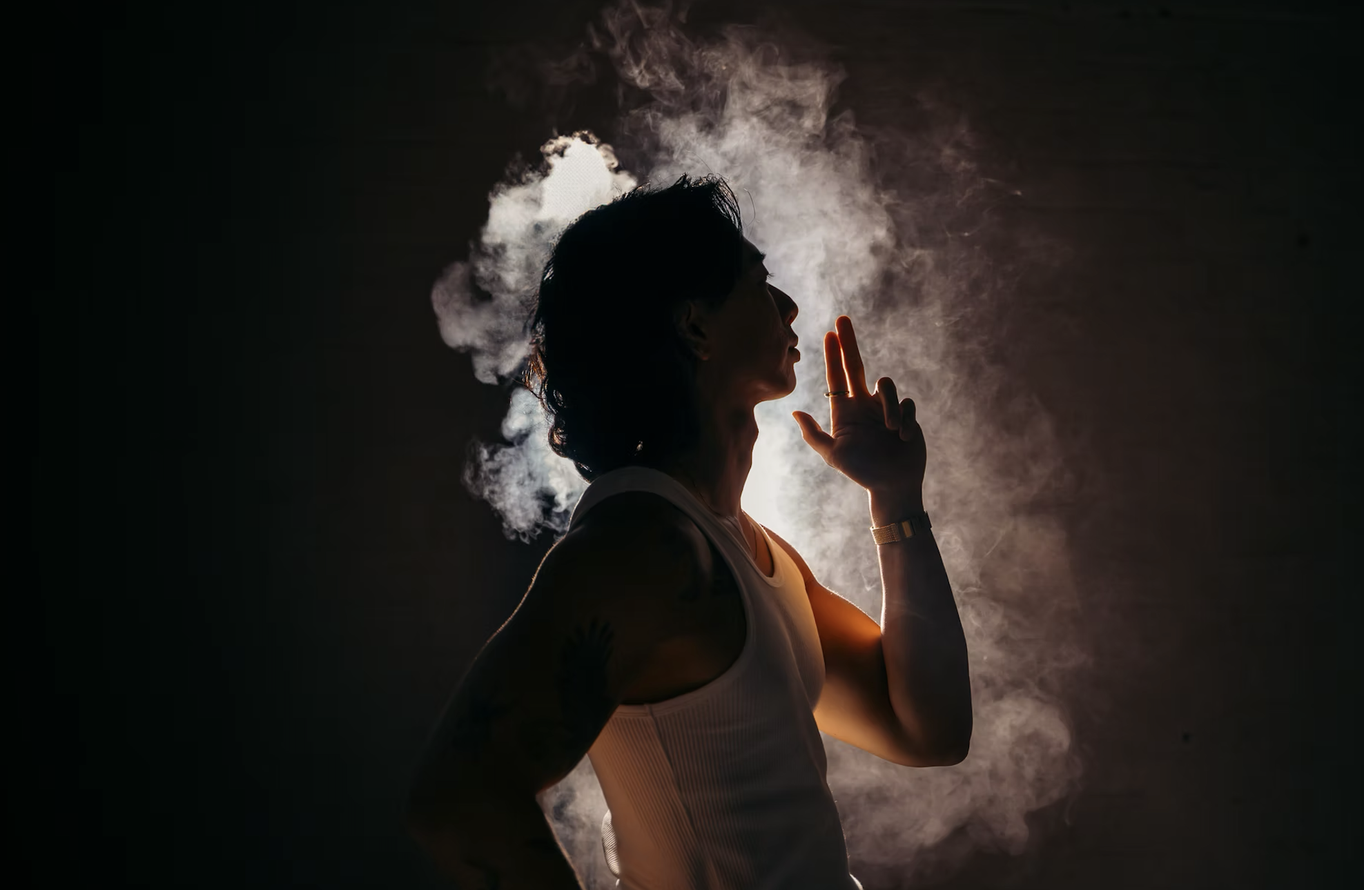 A woman with dark hair, wearing a sleeveless top, stands in profile against a dark background, with smoke illuminated by backlighting creating a halo effect around her head. She has her hand near her face, and the scene has a dramatic, mysterious atm