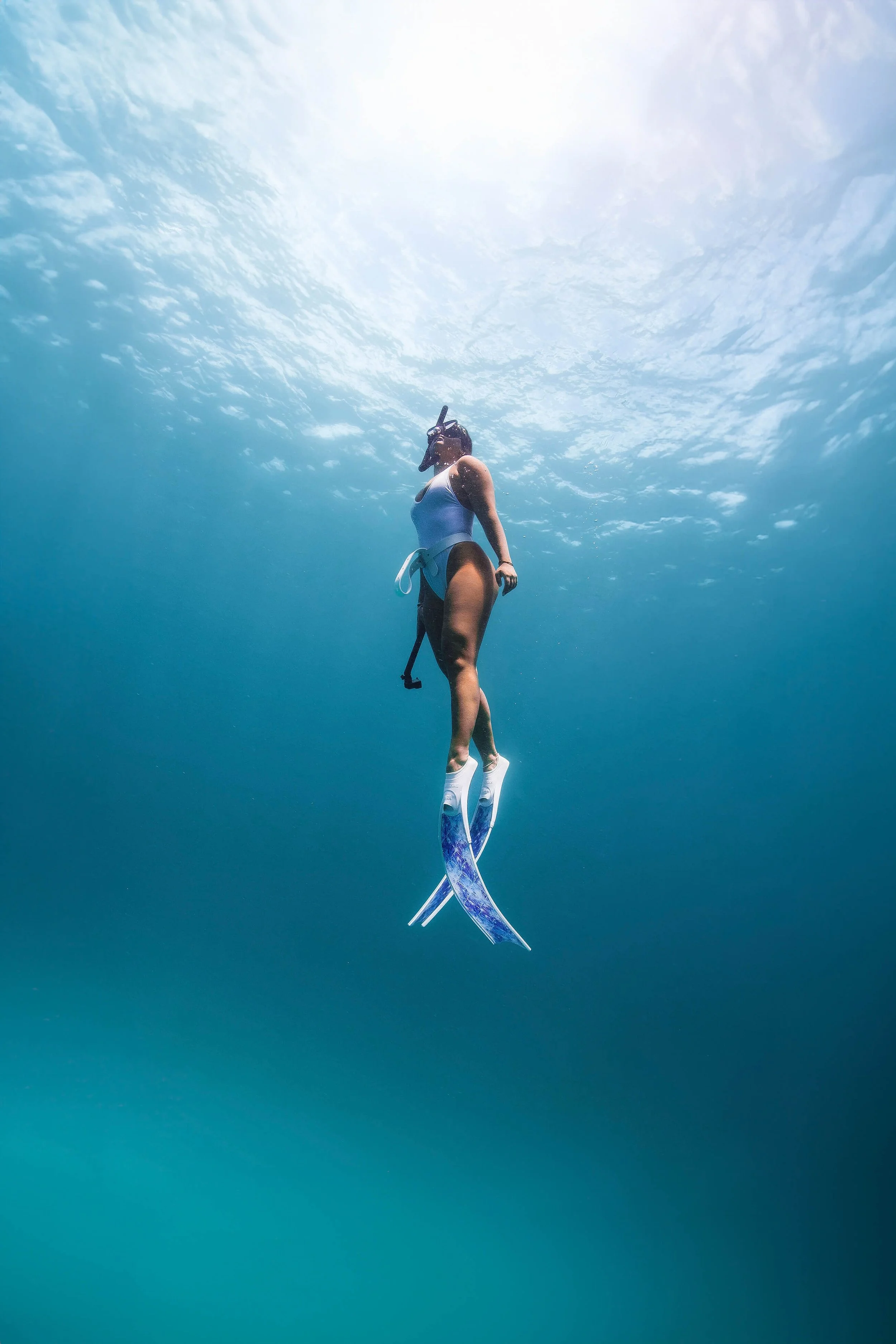 A person scuba diving underwater, surrounded by blue water and sunlight filtering from above, wearing a snorkel mask, a wetsuit top, flexible fins, and holding a snorkel.