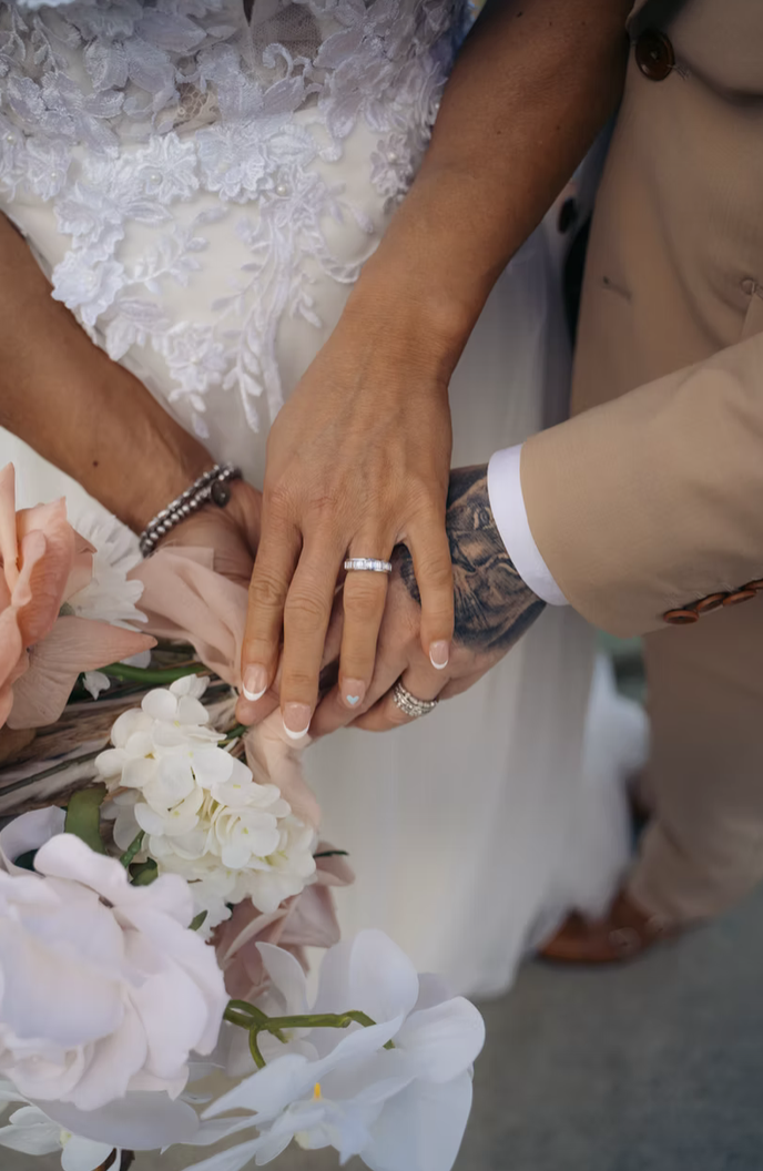Close-up of a bride and groom holding hands, displaying wedding rings, with a bouquet of white and pale pink flowers. The bride wears lace, and the groom has a tattooed arm.