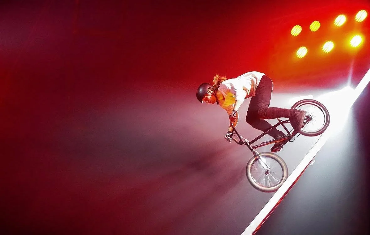 A person performing a trick on a BMX bike on an indoor ramp, with red and white lighting and yellow spotlights overhead.