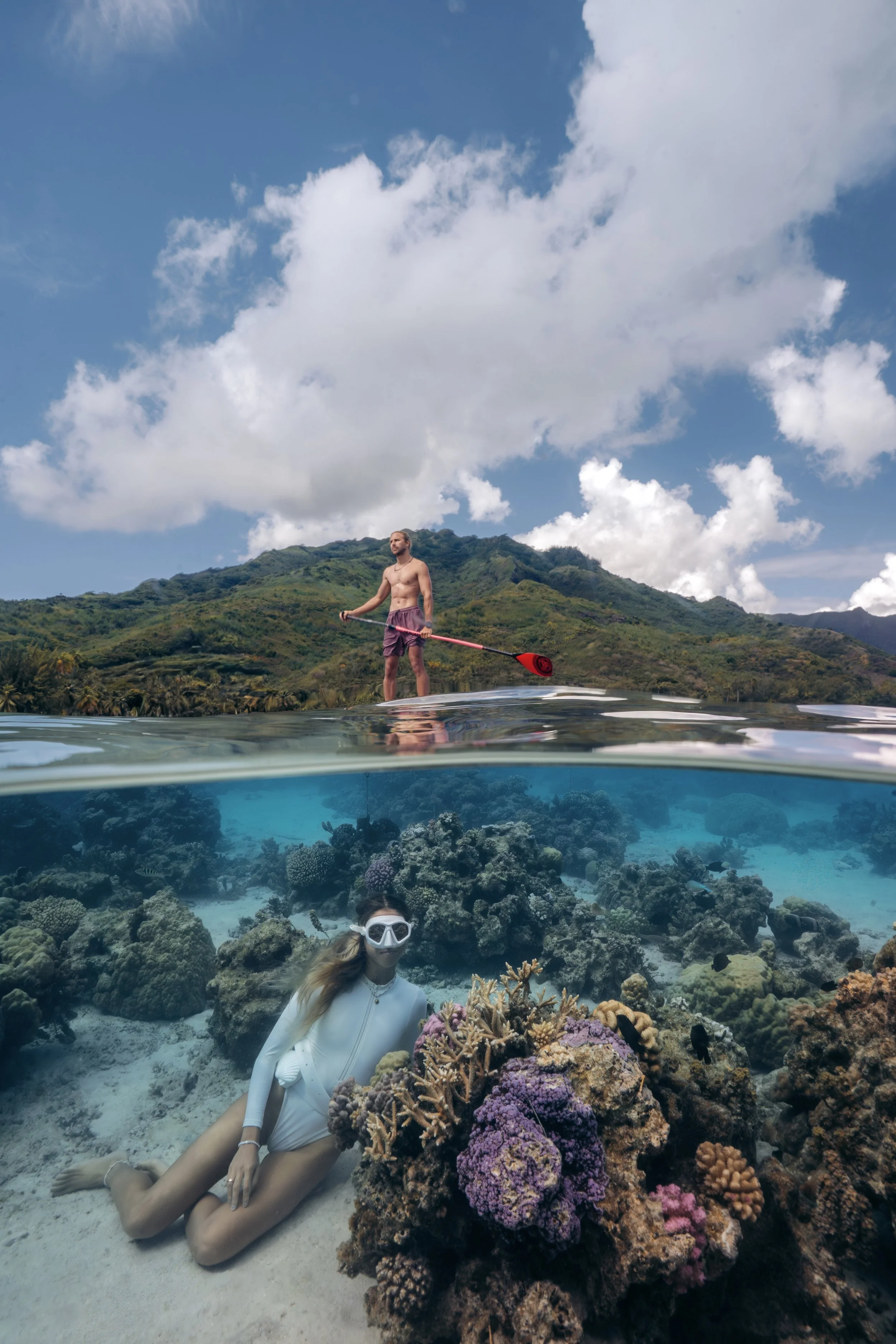 This image captures a split view: the top shows a man in shorts paddleboarding on open water with green hills in the background and a partly cloudy sky; the bottom shows a woman underwater near coral reefs, wearing a mask and swimsuit, with vibrant p
