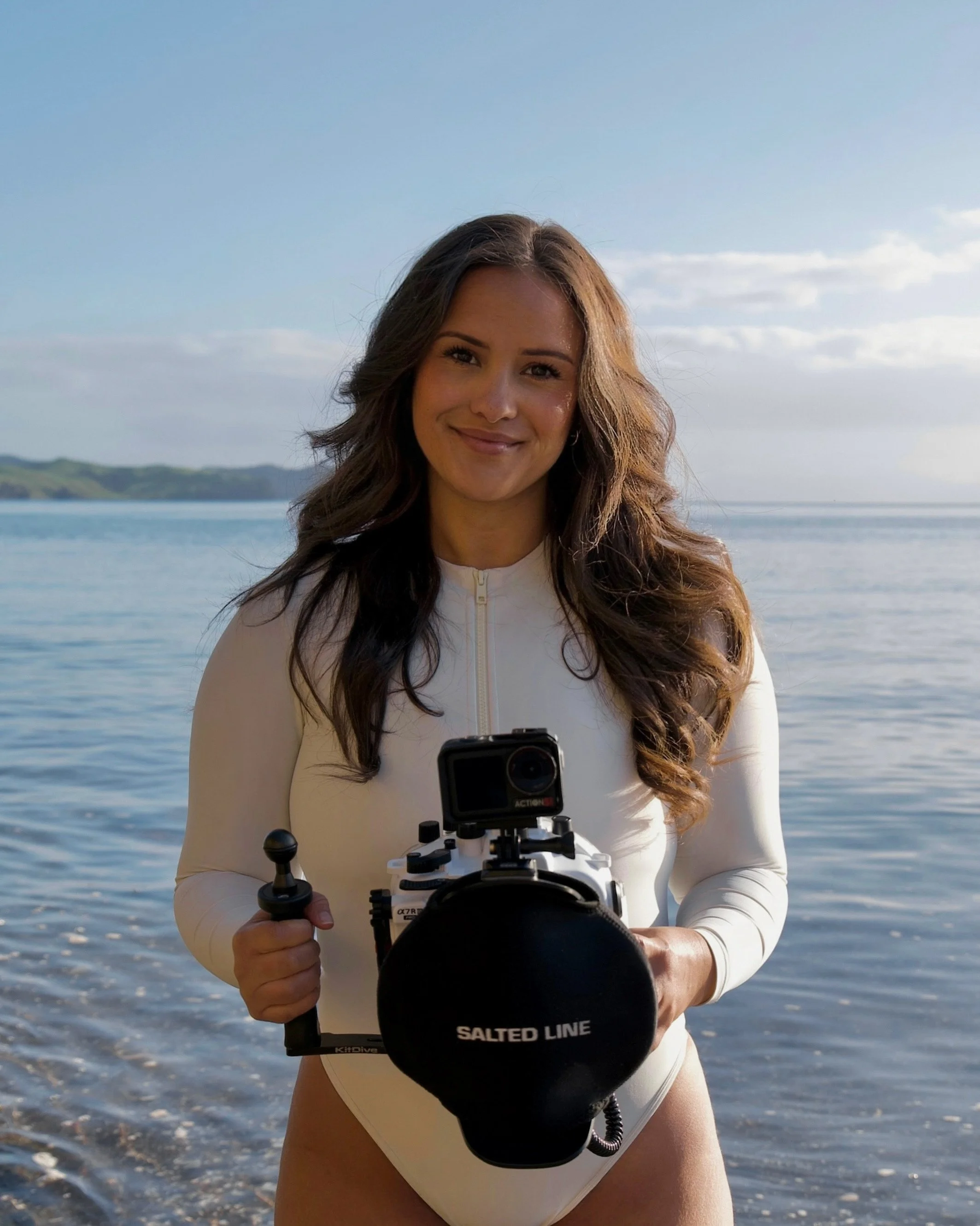 Woman in white outfit standing in front of a body of water holding a device with a camera and a large microphone.