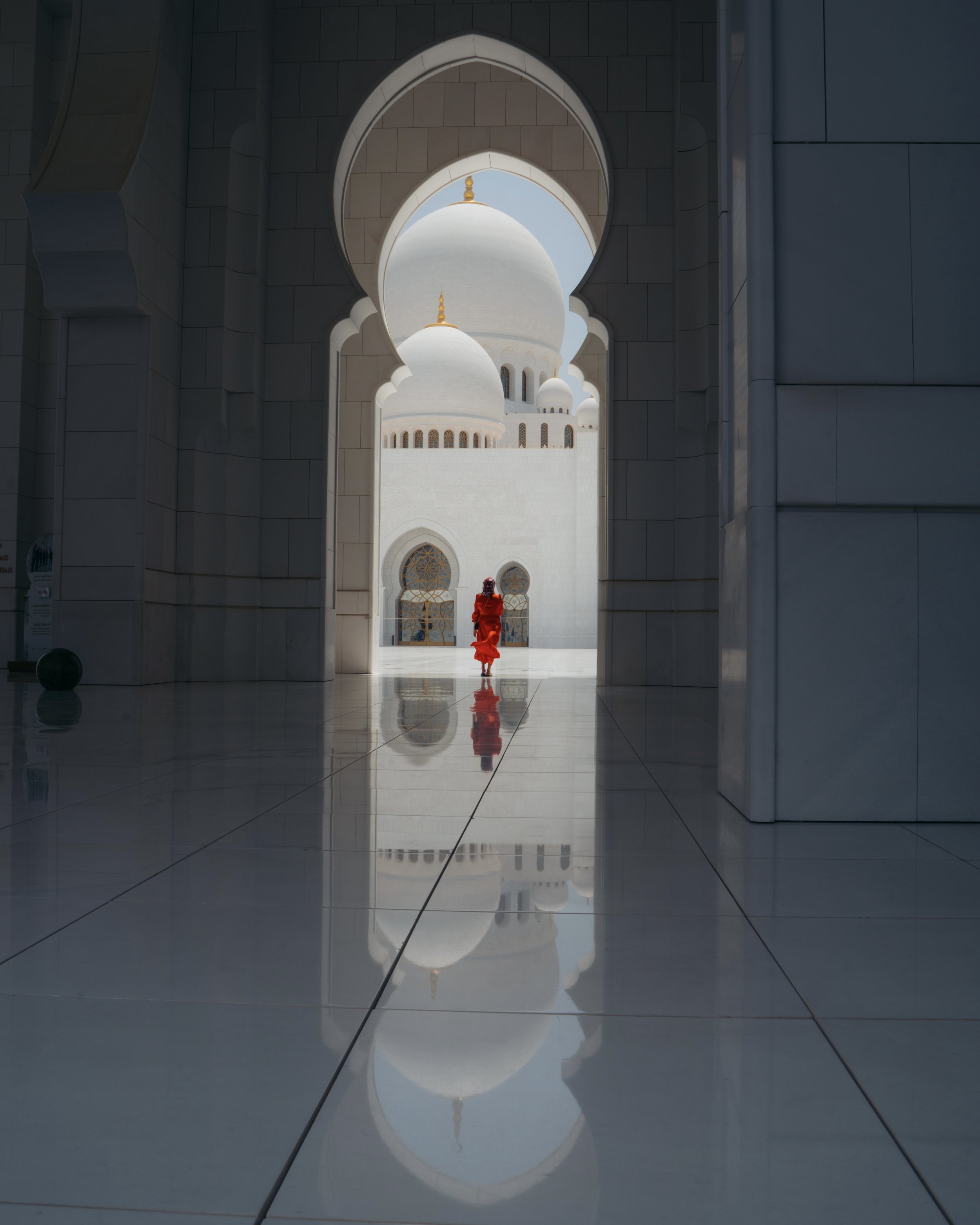 View through an arched opening to the white domes and minarets of a mosque, with a woman in a red dress walking in the courtyard, and her reflection on the polished floor.