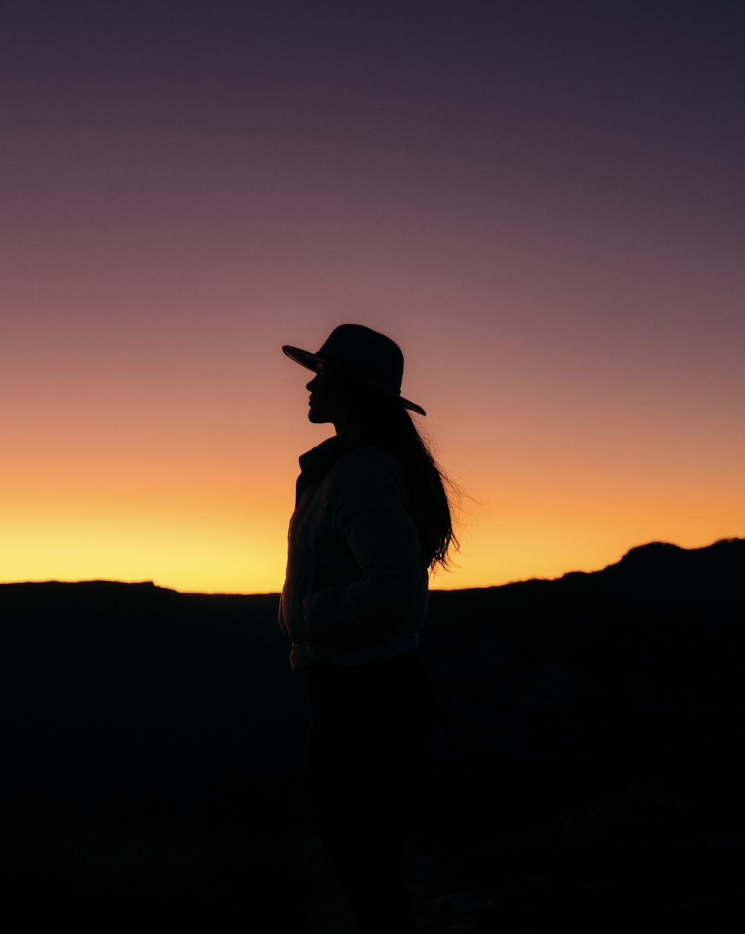 Silhouette of a woman with a hat standing in front of a colorful sunset sky.
