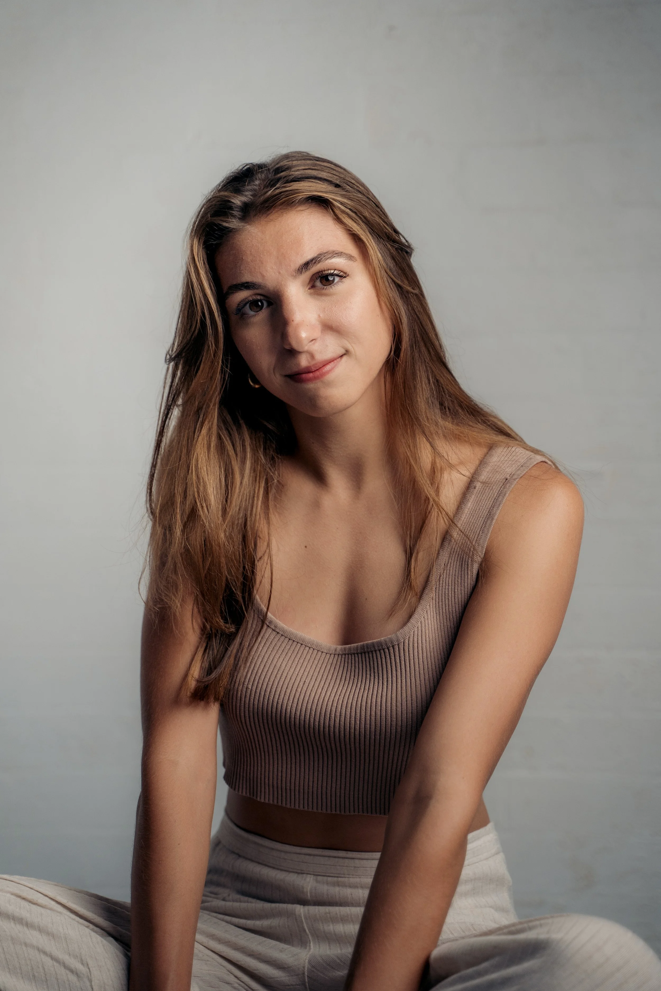 Young woman with long wavy brown hair wearing a beige sleeveless ribbed top and beige pants, sitting cross-legged against a plain gray background.