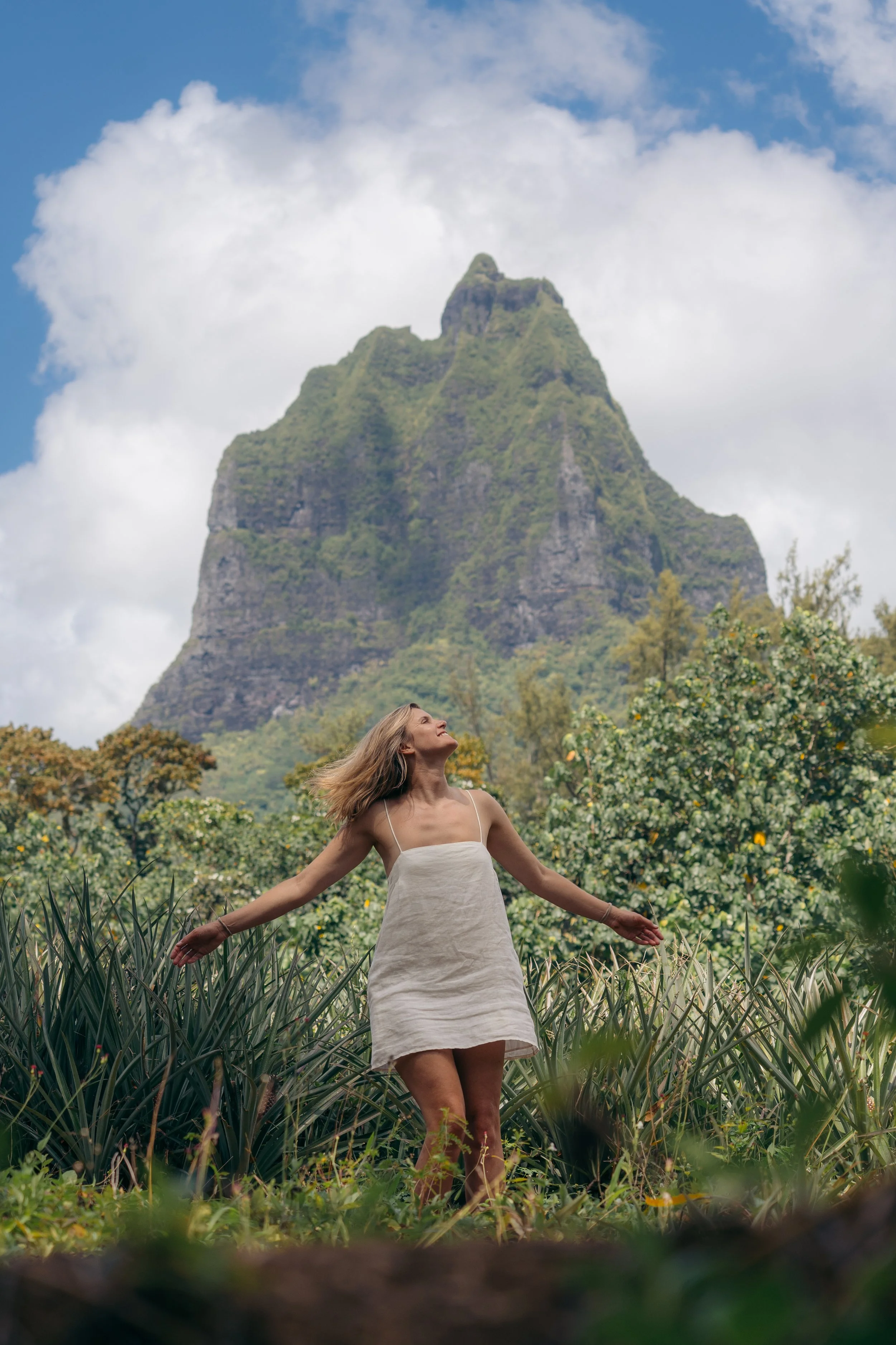 A woman in a white dress with her arms outstretched standing in a green field of tall grass or crops, with a large rocky mountain covered in greenery in the background, under a partly cloudy sky.