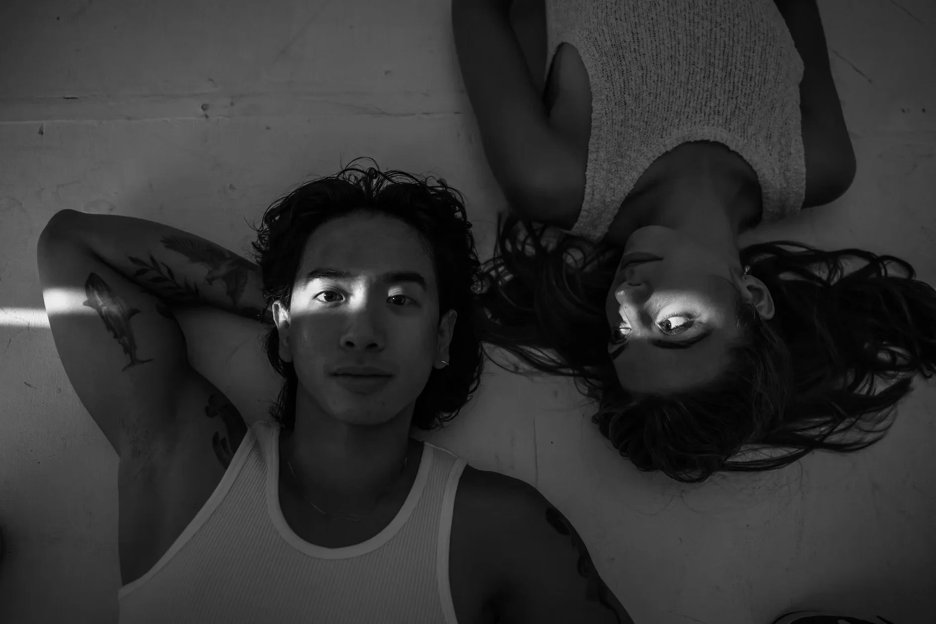 A black and white photo of a young man and woman lying on the floor side by side. The man has short, curly hair, tattoos on his arm, and wears a sleeveless top. The woman has long wavy hair and wears a sleeveless top. They are looking up at the camer
