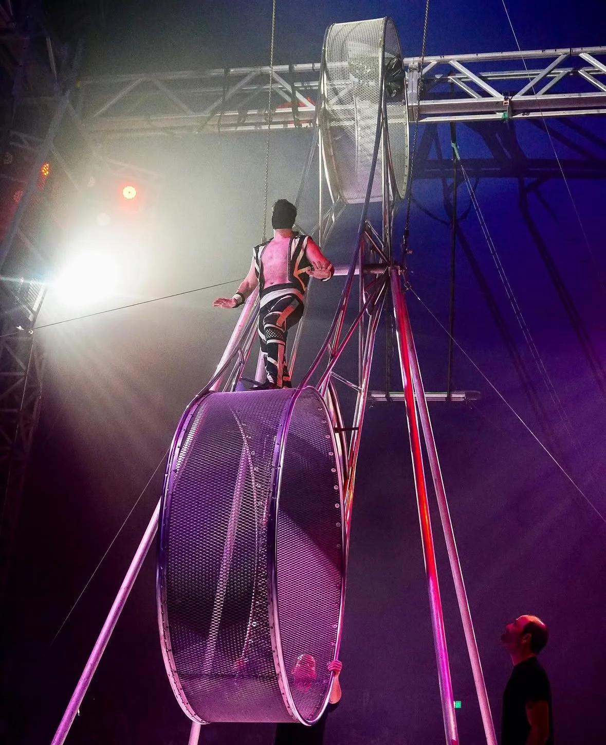 A performer is walking on a large metal cylinder on top of a high scaffold in a circus tent, illuminated by stage lights and smoke.