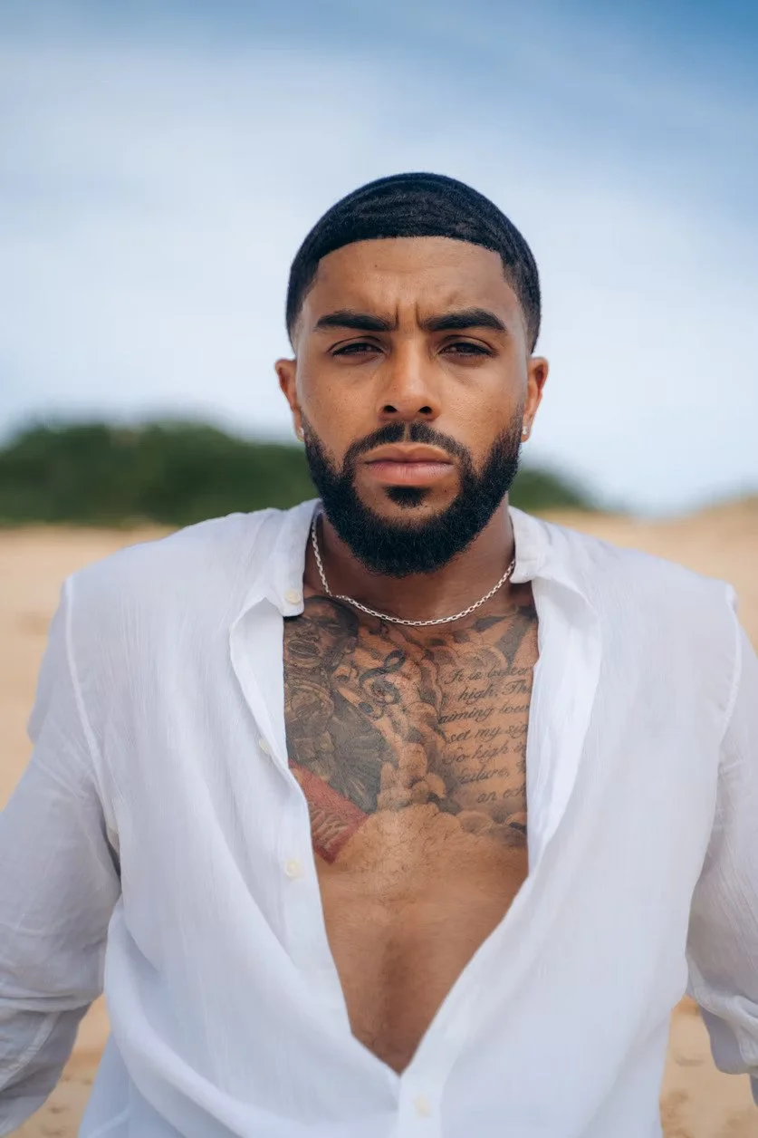 A man with a beard and short hair, wearing a white open shirt, standing on a beach with sand and greenery in the background.