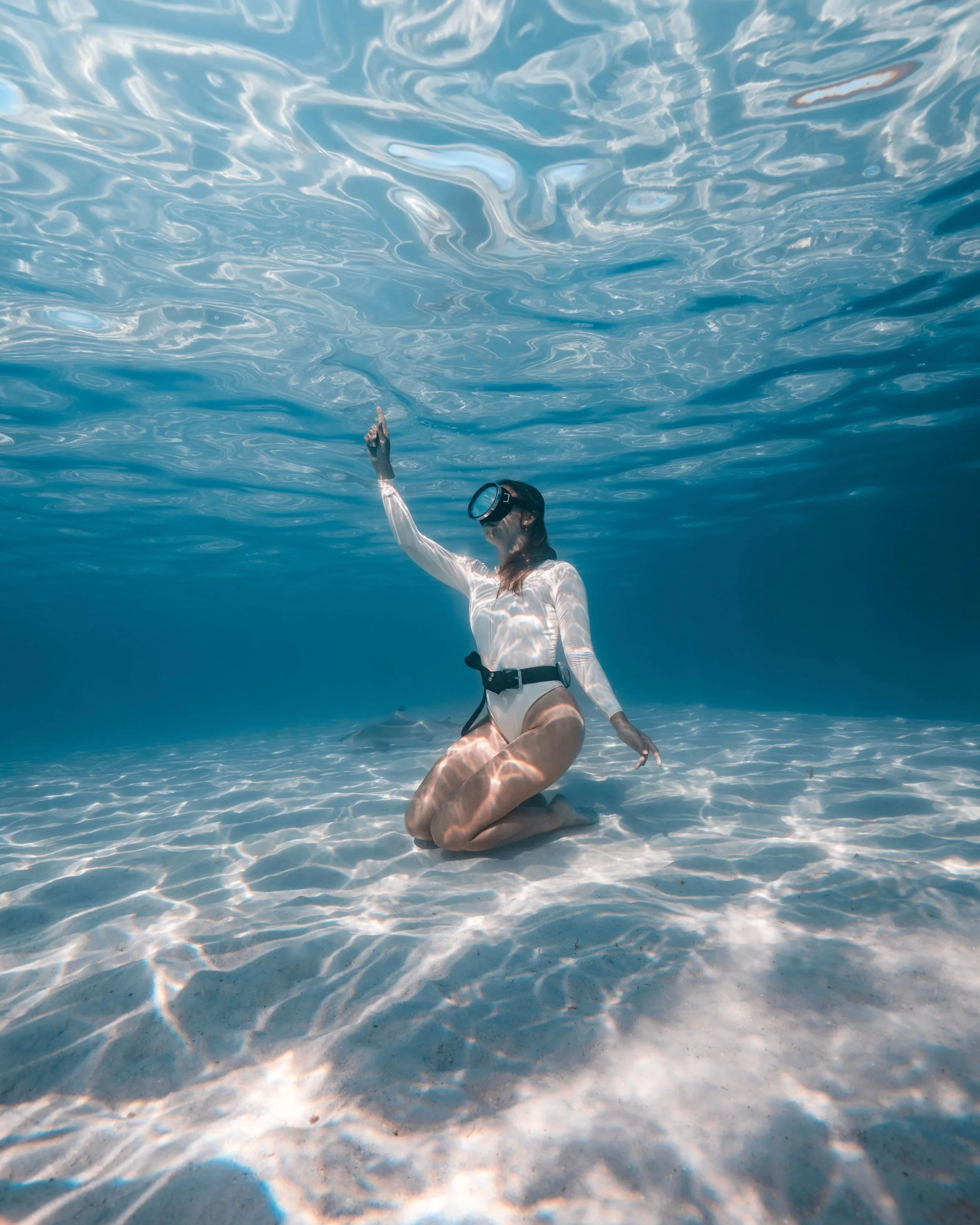 A person wearing a wetsuit and snorkeling gear kneels on the sand underwater in the ocean, reaching up towards the water surface with rippling blue water around.