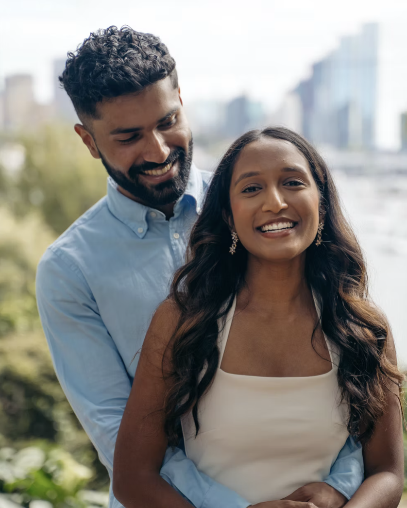 A smiling couple outdoors with city buildings in the background, the man standing behind the woman, both wearing casual attire.