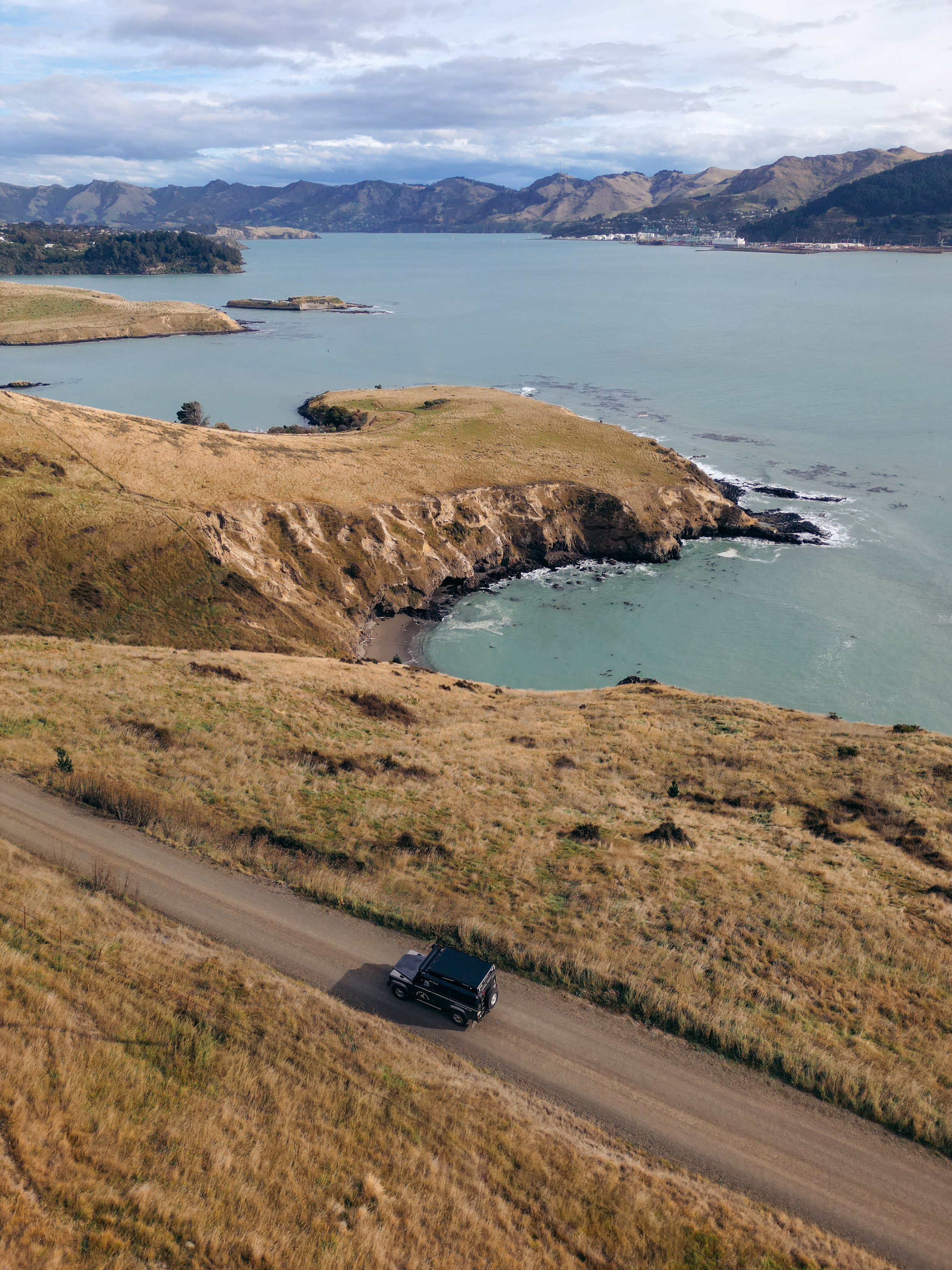 A black off-road vehicle driving on a dirt road through a grassy hillside with a large body of water and mountainous landscape in the background.