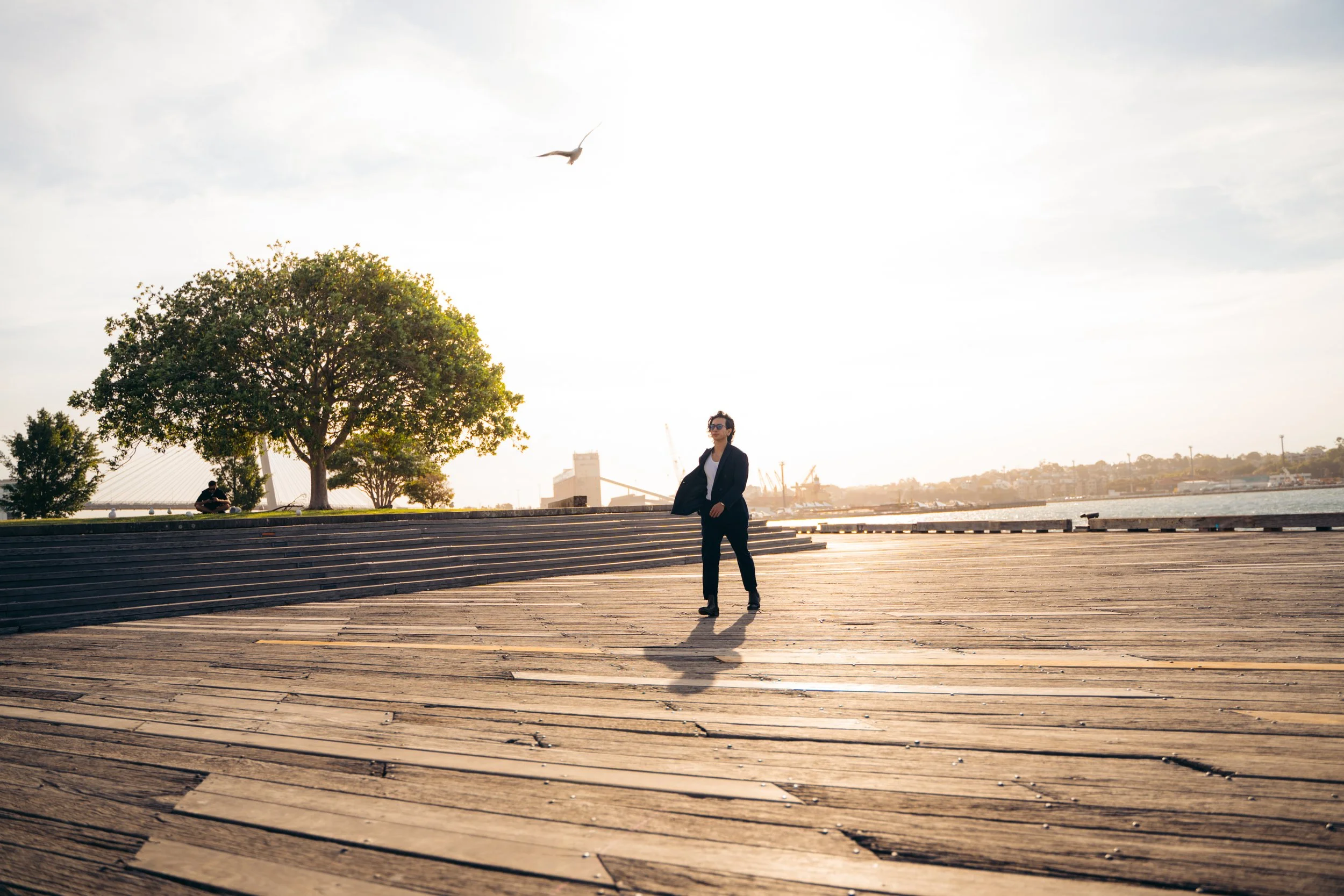 A woman with dark hair and glasses standing on a wooden boardwalk near a waterfront, with trees, buildings, and a bridge in the background during sunset.