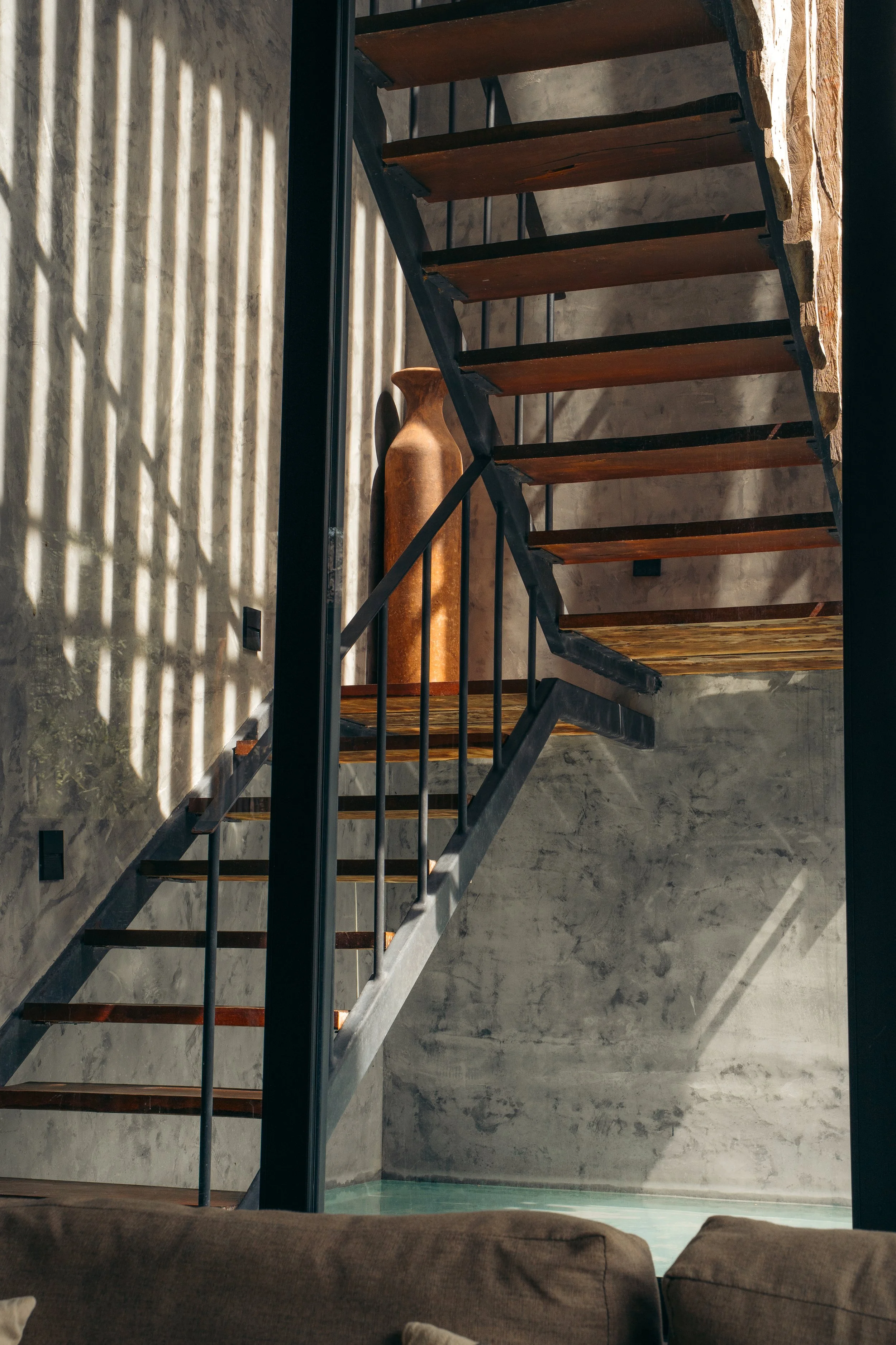 Interior view of a staircase with wooden steps, metal railing, and concrete walls. Sunlight creates striped shadows on the wall, and a decorative wooden vase is placed on the landing.