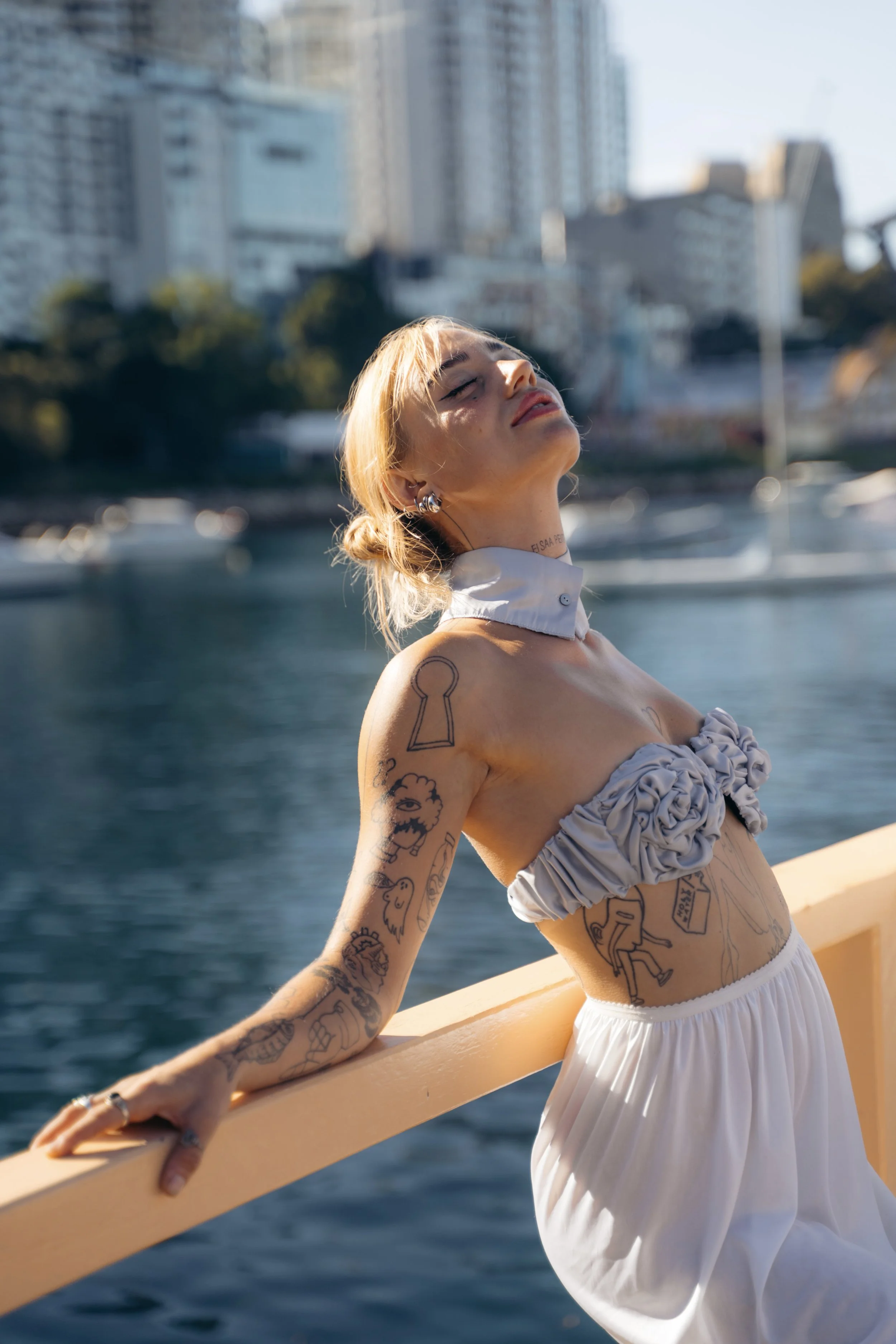 A woman with blonde hair, tattoos on her arms and chest, and earrings is standing on a boat with water and city buildings in the background. She is smiling with her eyes closed, wearing a ruffled top and a white skirt, enjoying a sunny day.