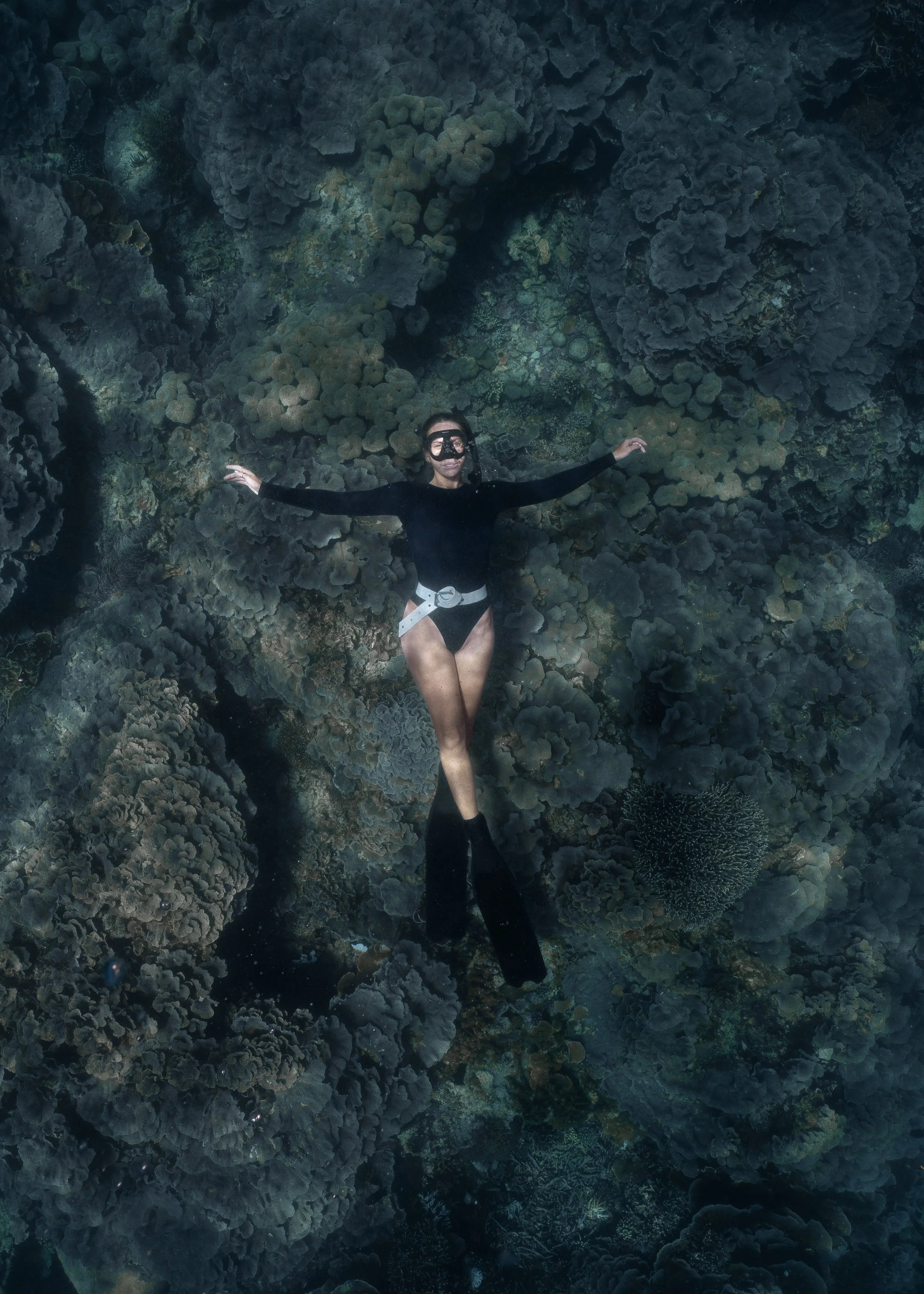 Person snorkeling underwater among coral reef, wearing black wetsuit, mask, and snorkel, with arms outstretched.