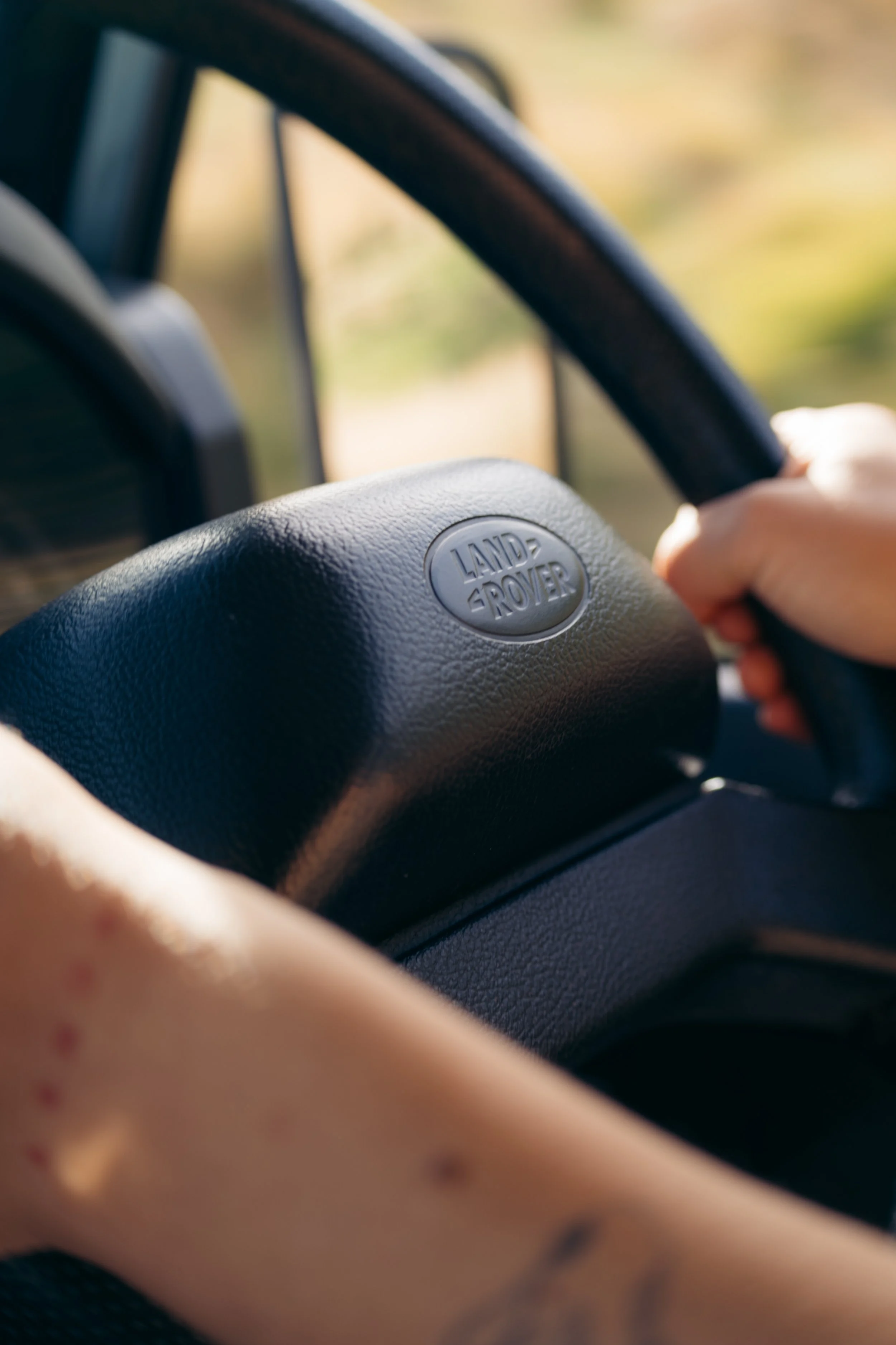 Close-up of a Land Rover steering wheel being held by a person inside a vehicle, with a blurred outdoor background.