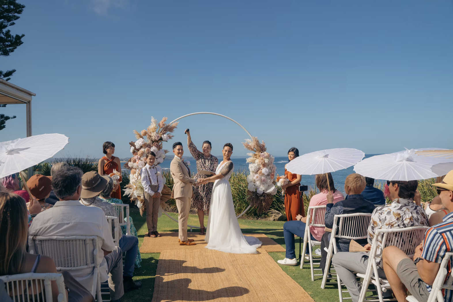 A wedding ceremony outdoors by the ocean with guests sitting under umbrellas, the couple exchanging vows, and a floral arch in the background.