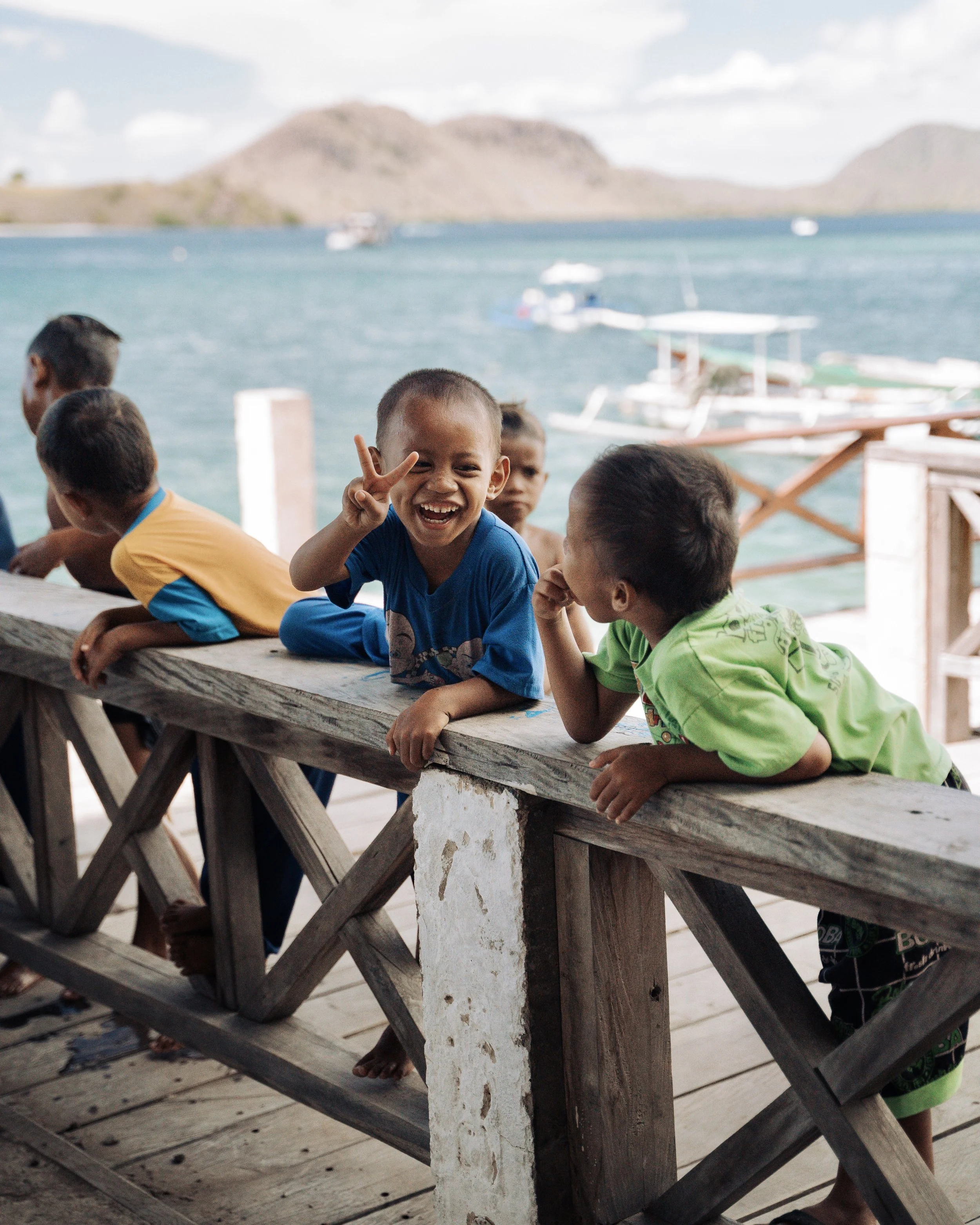 Group of children sitting by a wooden railing near a body of water with boats and mountains in the background. One child is smiling and making a peace sign.