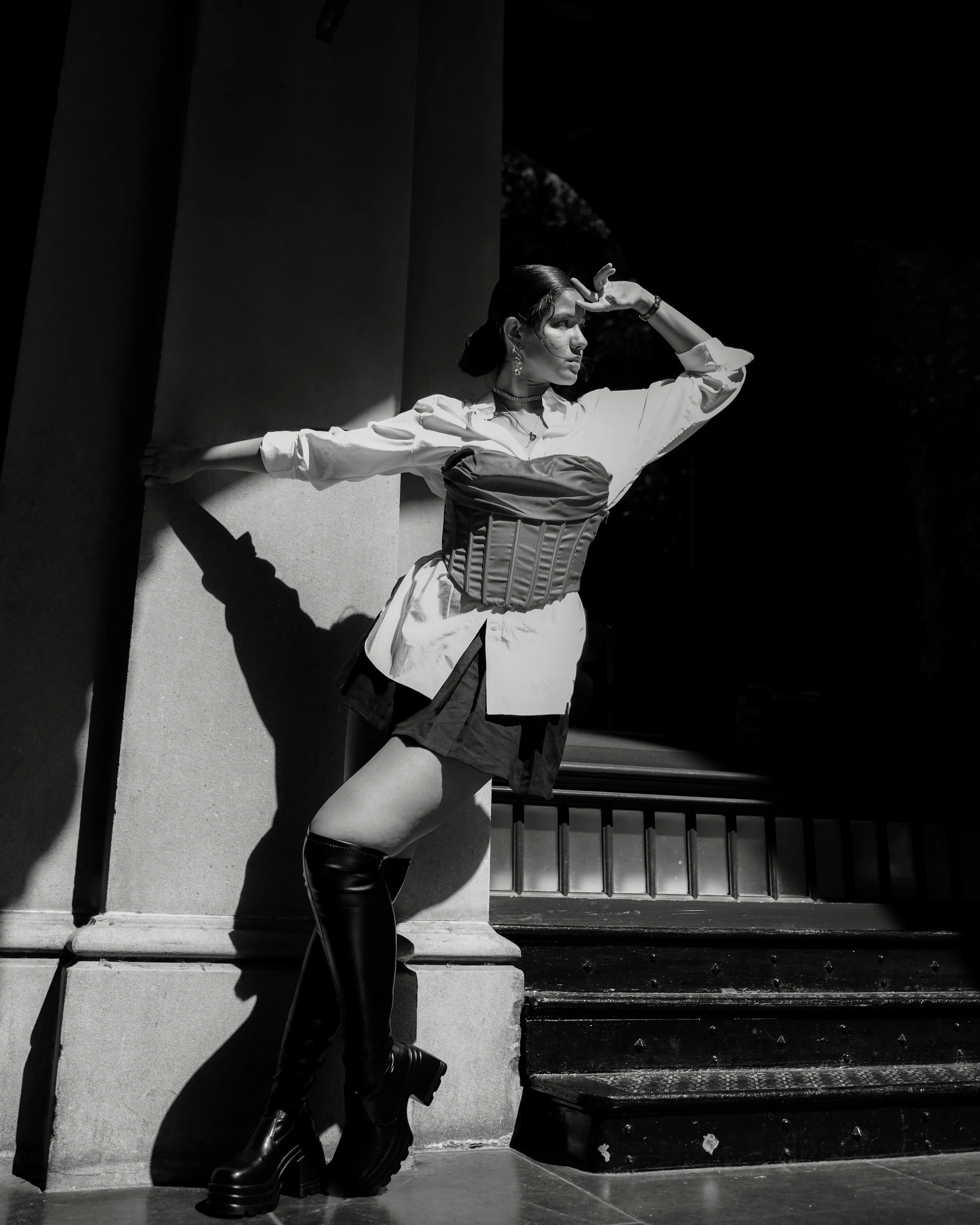 A woman standing on a staircase in sunlight, dressed in a corset, blouse, and thigh-high boots, with one hand on her forehead and the other resting on the wall.