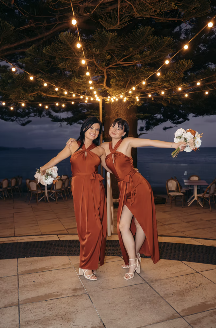 Two women in matching rust-colored dresses dancing under string lights on an outdoor patio near the beach at dusk, holding bouquets.