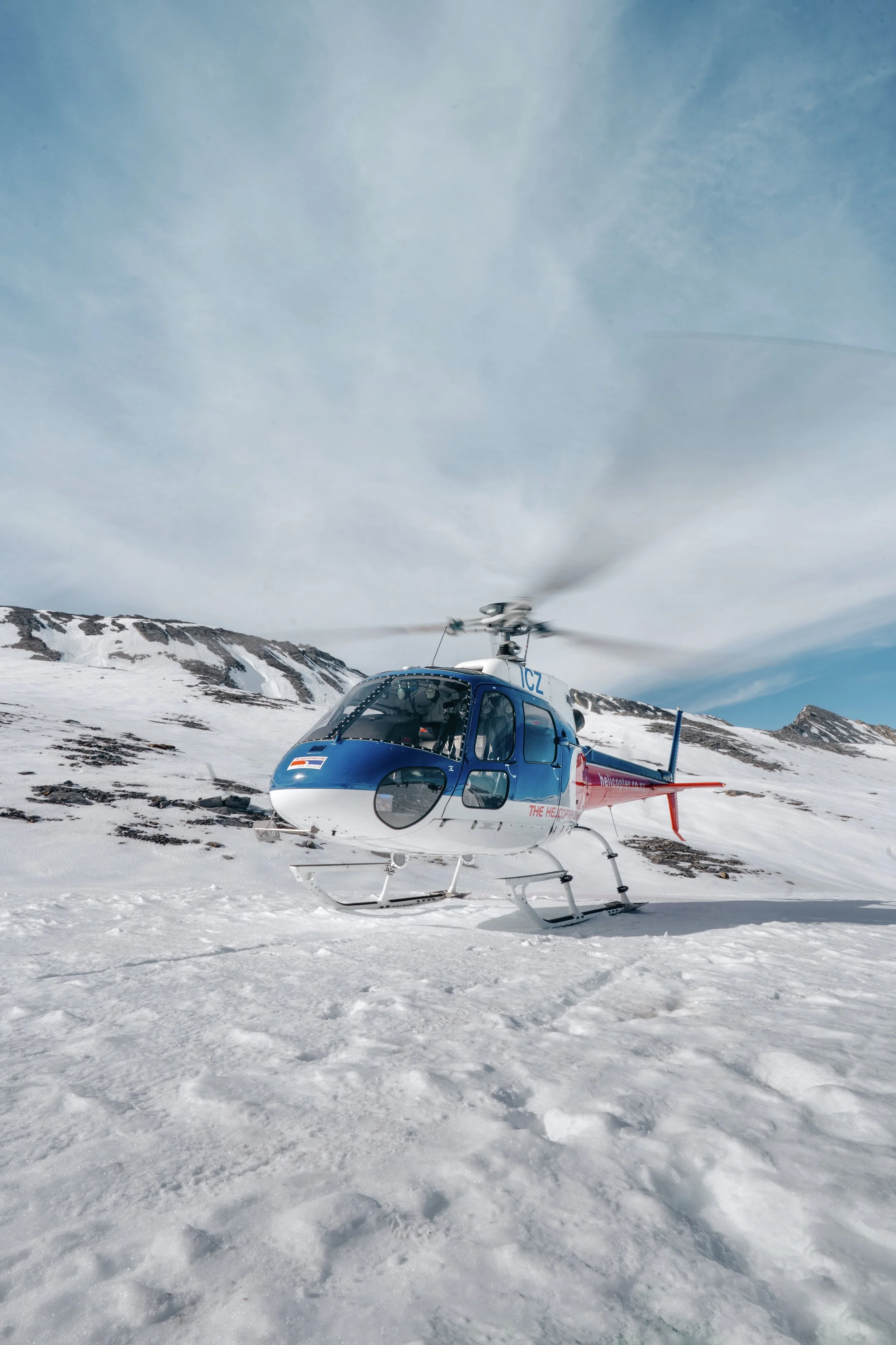 A helicopter on snowy terrain with mountains in the background and a partly cloudy sky above.